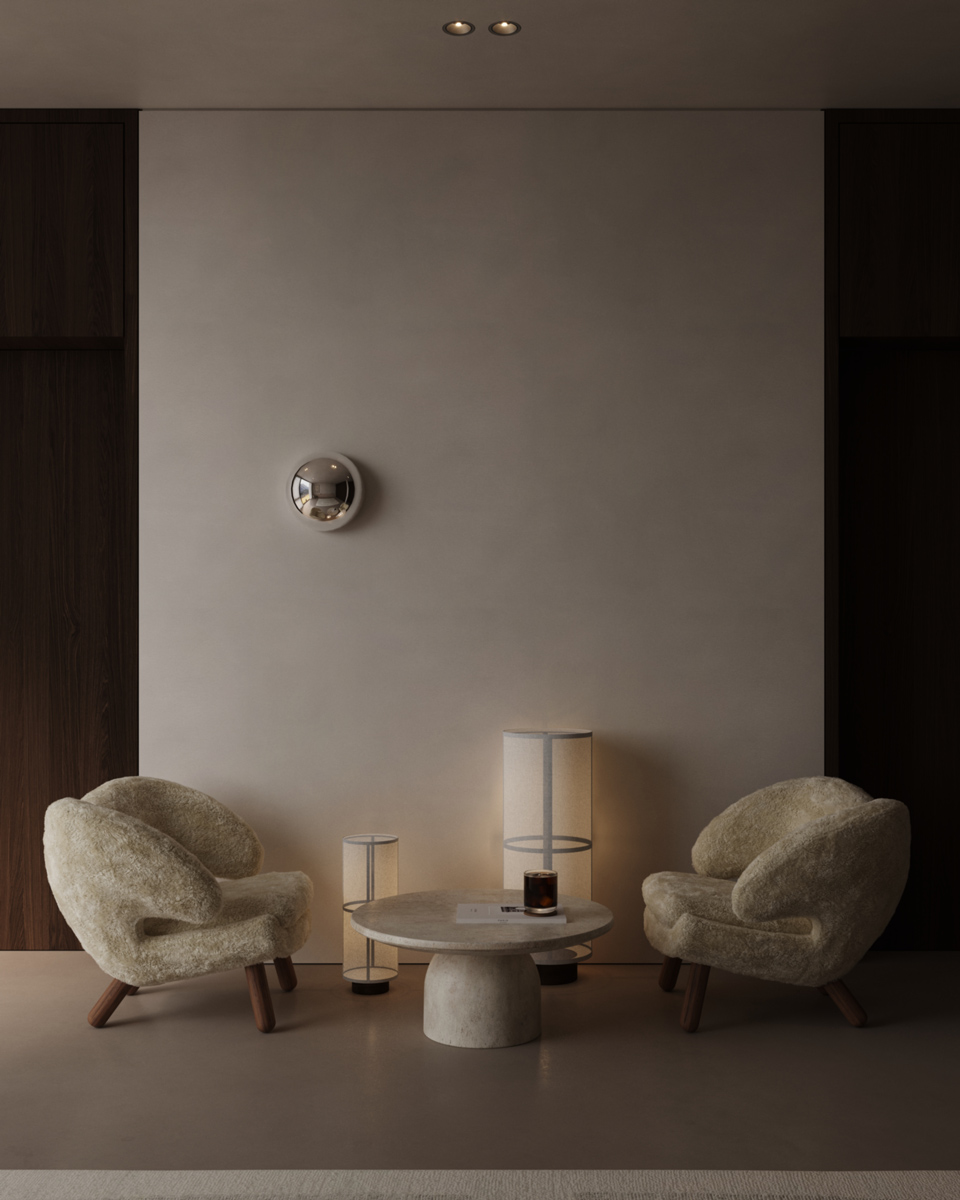 Minimalist interior seating area with two cream-colored fuzzy armchairs facing each other, a round stone coffee table between them holding a dark glass candle and a booklet, and two cylindrical floor lamps emitting warm light against a beige wall accented by dark wooden panels on both sides and a small reflective metallic wall decor piece.