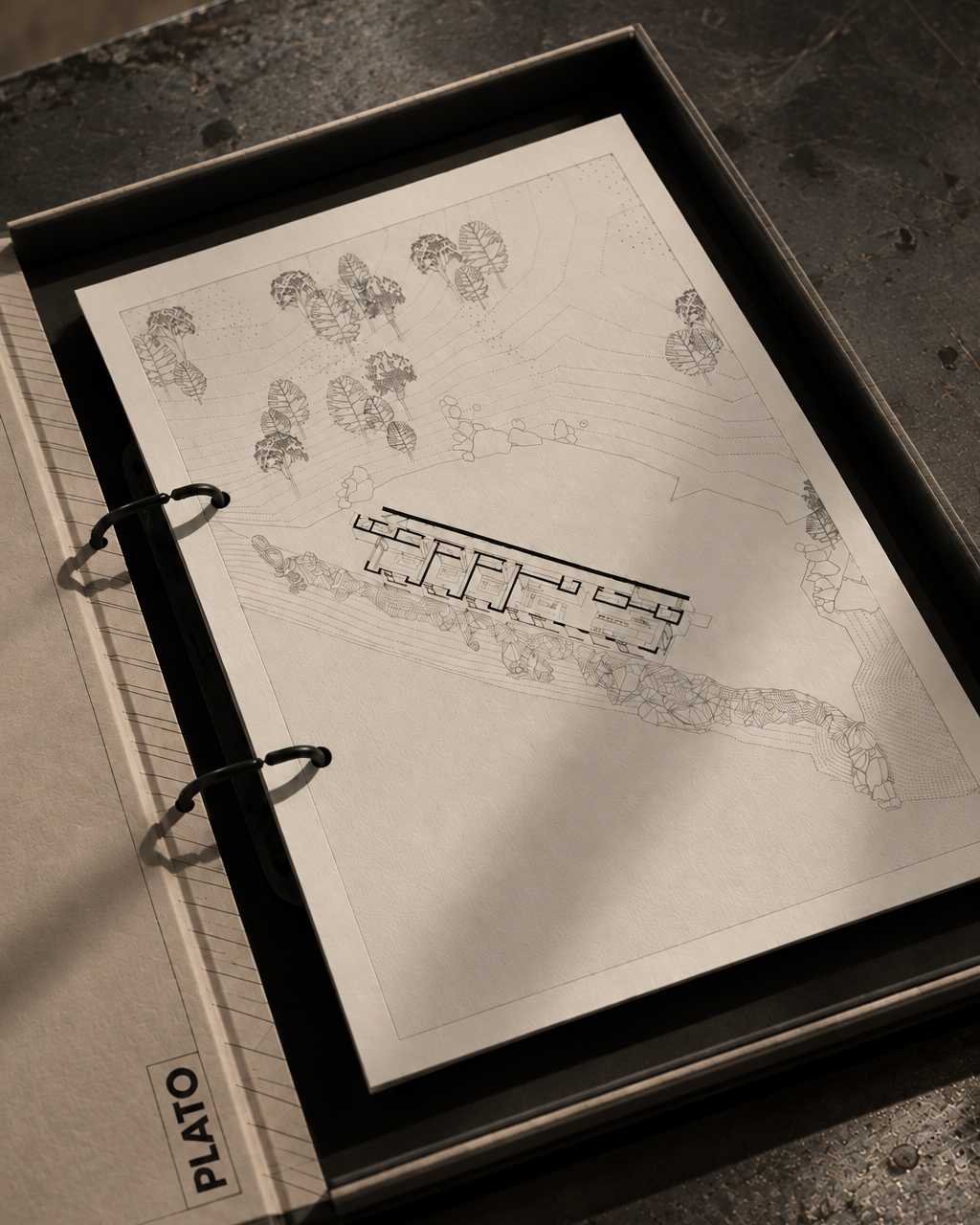 Architectural floor plan of a building with interior rooms and surrounding landscape depicted with trees and terrain contour lines, held in a binder labeled 'PLATO' on a textured surface.