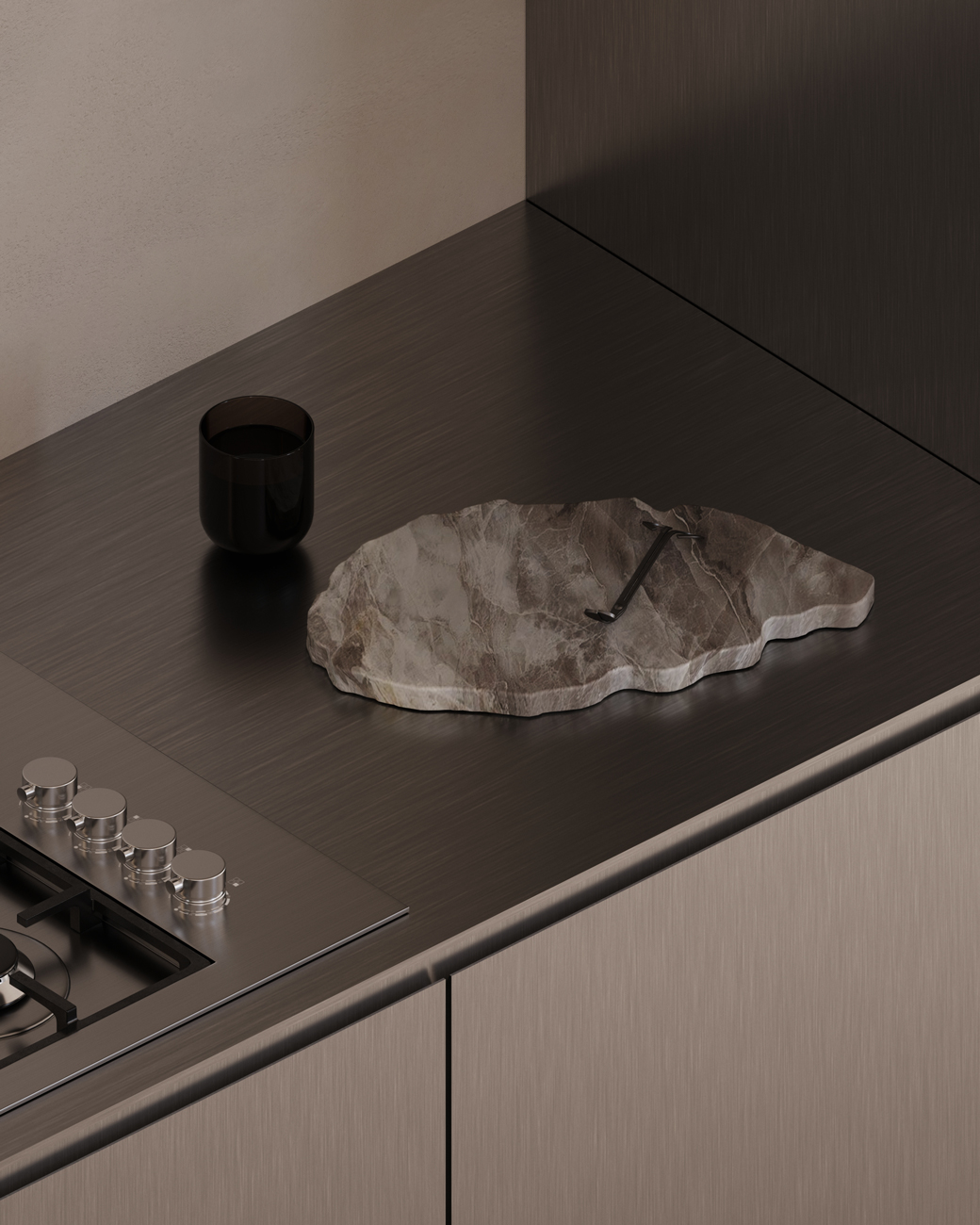 Modern kitchen countertop with dark brushed metal surface, a black glass cup, and an irregularly shaped marble cutting board with a metal tool resting on it, next to a stainless steel gas stove with control knobs.