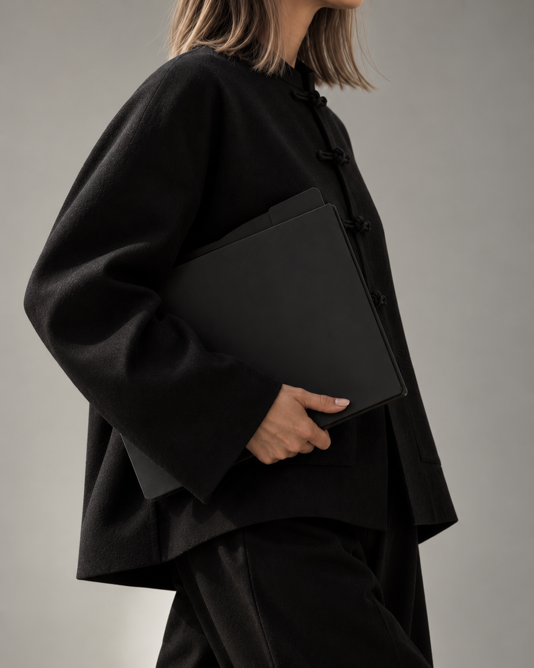 Person wearing a black traditional-style jacket with frog closures holding a closed black laptop against their side, against a neutral gray background.