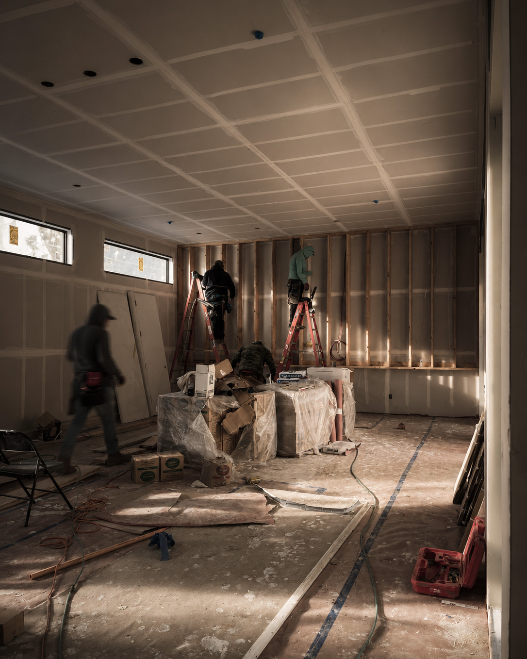 Interior of a room under construction with unfinished walls and ceiling, workers on ladders installing or finishing wall framing, and construction materials scattered on the floor and covered with plastic.