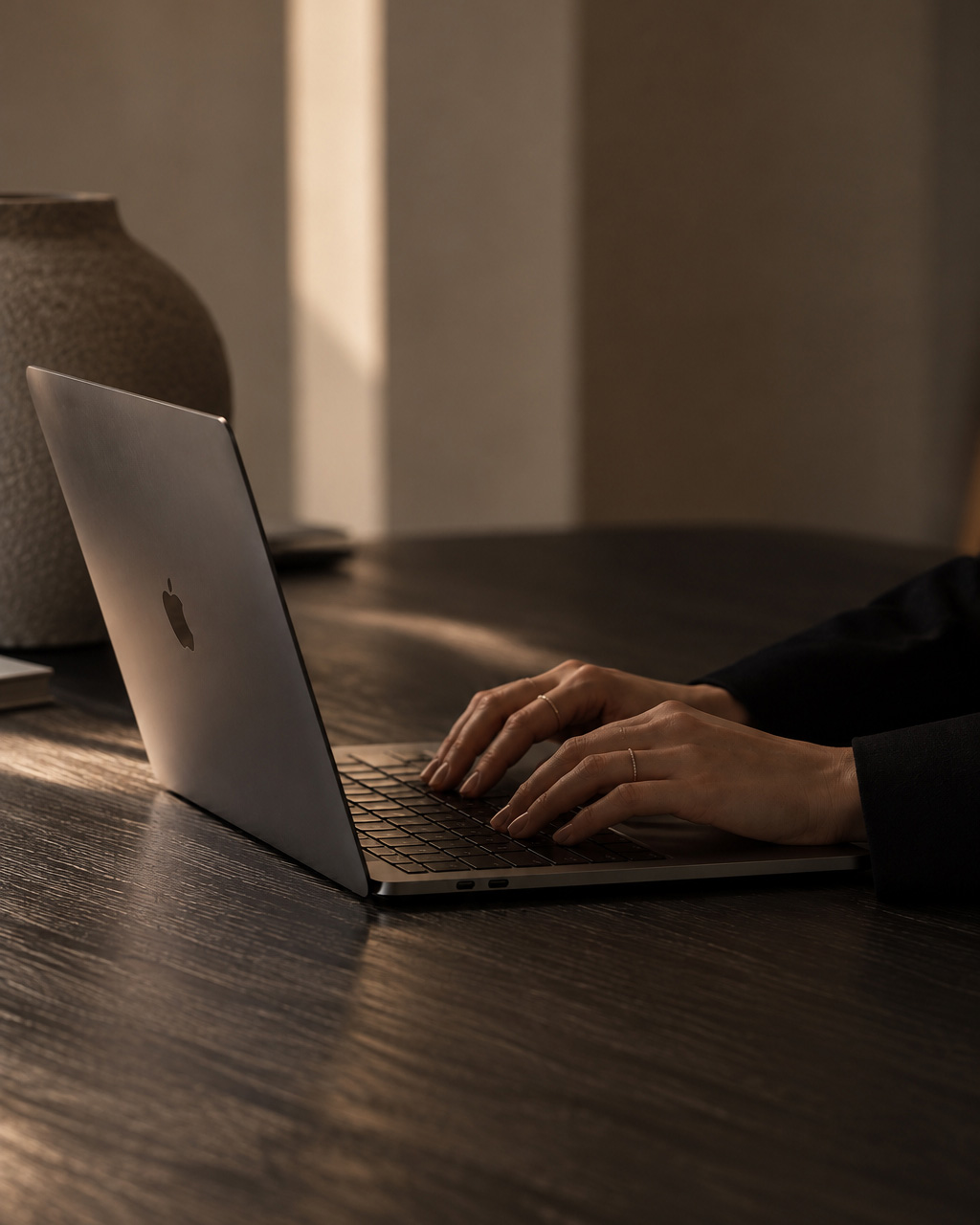 Close-up of hands typing on a MacBook laptop keyboard on a dark wooden table with soft warm lighting and a textured vase in the background.