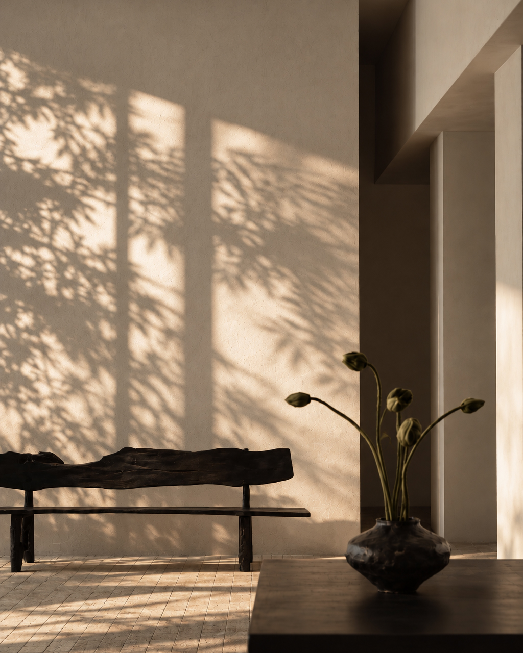 Sunlight casts shadows of leaves on a beige textured wall behind a dark rustic wooden bench, with a dark vase holding budding flowers on a table in the foreground.