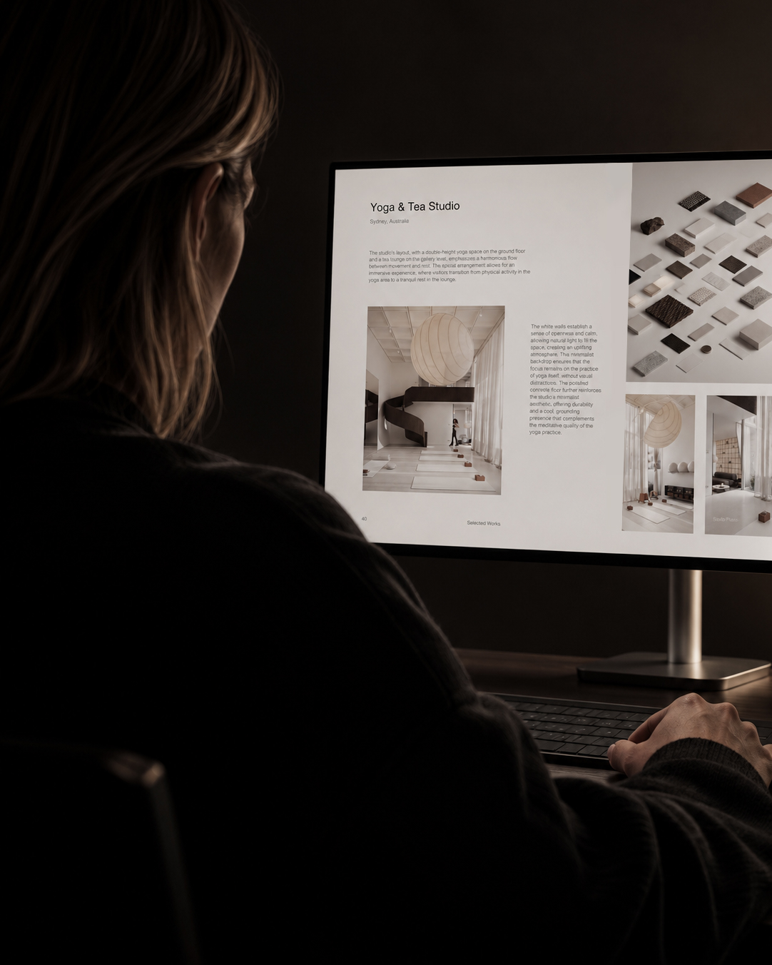 Person viewed from behind looking at a computer monitor displaying a webpage titled 'Yoga & Tea Studio' with images and text describing a yoga studio design featuring a large hanging light, spiral staircase, and a display of material samples.