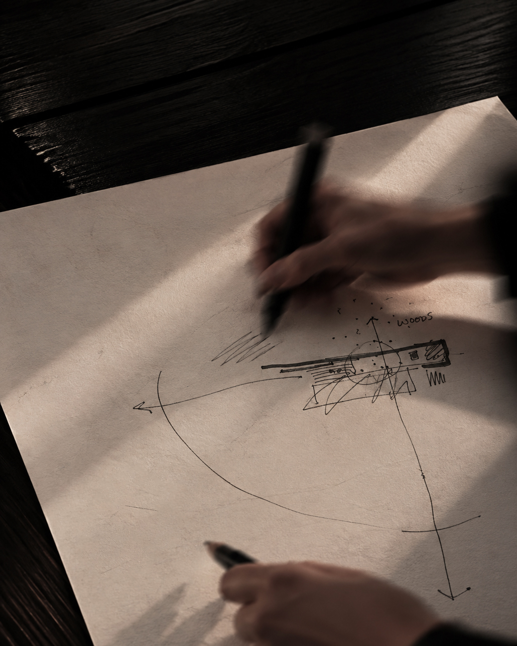 Person sketching a rough architectural or landscape plan with ink on a beige sheet of paper laid on a dark wooden table, showing arrows and the word 'woods'.
