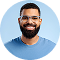 Smiling man with glasses and beard wearing a blue shirt against a blue background.