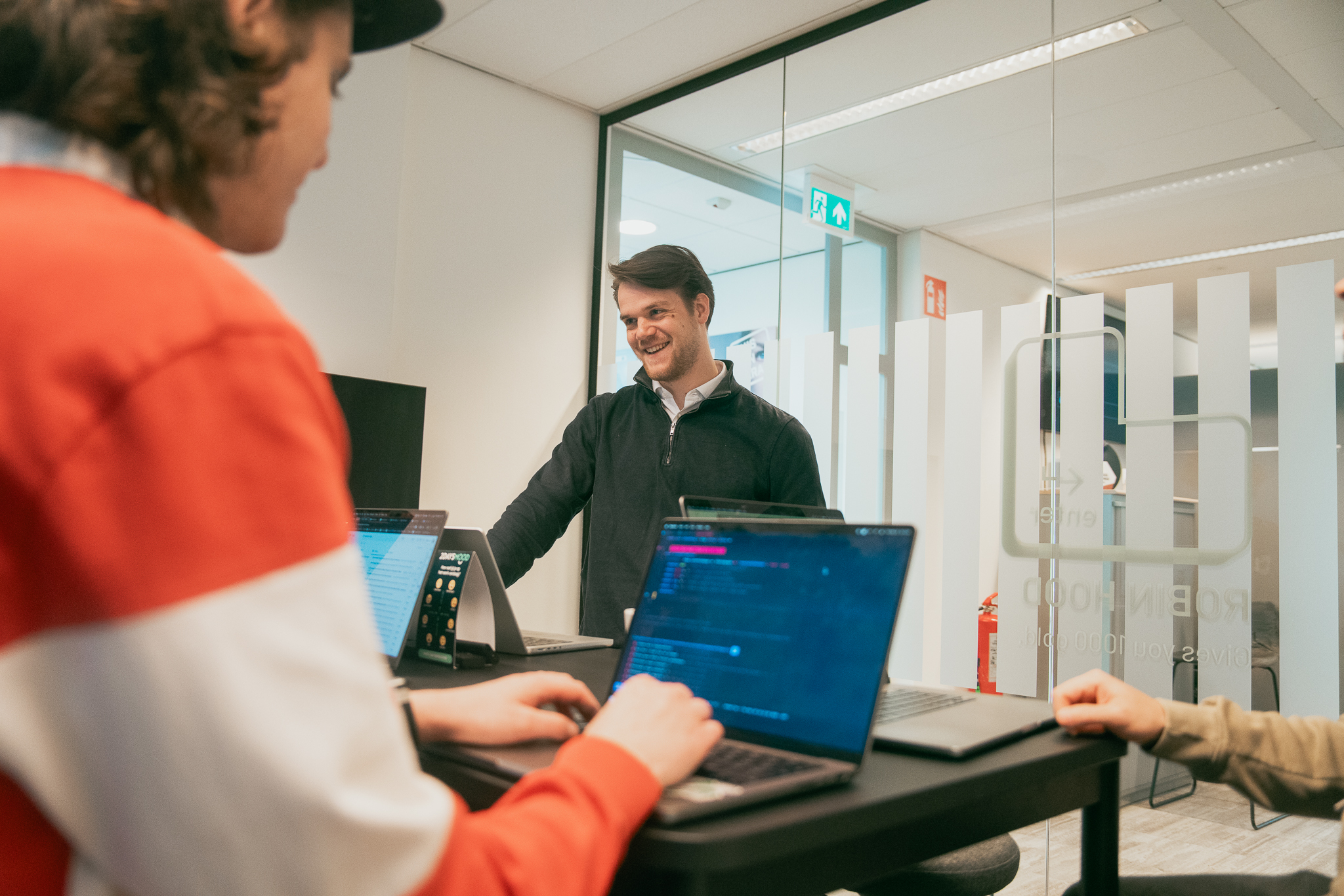 Man in black sweater smiling at two people working on laptops at a standing desk in an office.