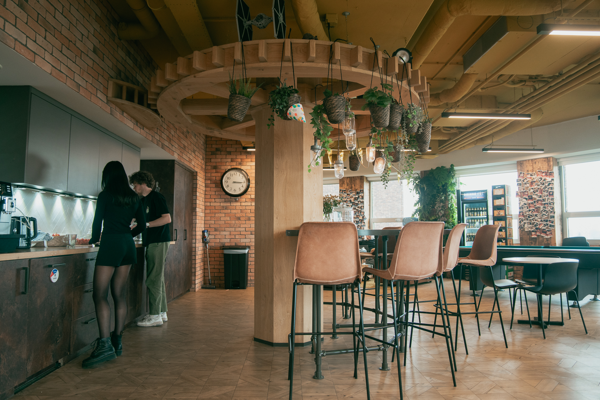 Modern lounge with hanging plants, tall tan chairs, brick walls, and two people near a kitchen counter.