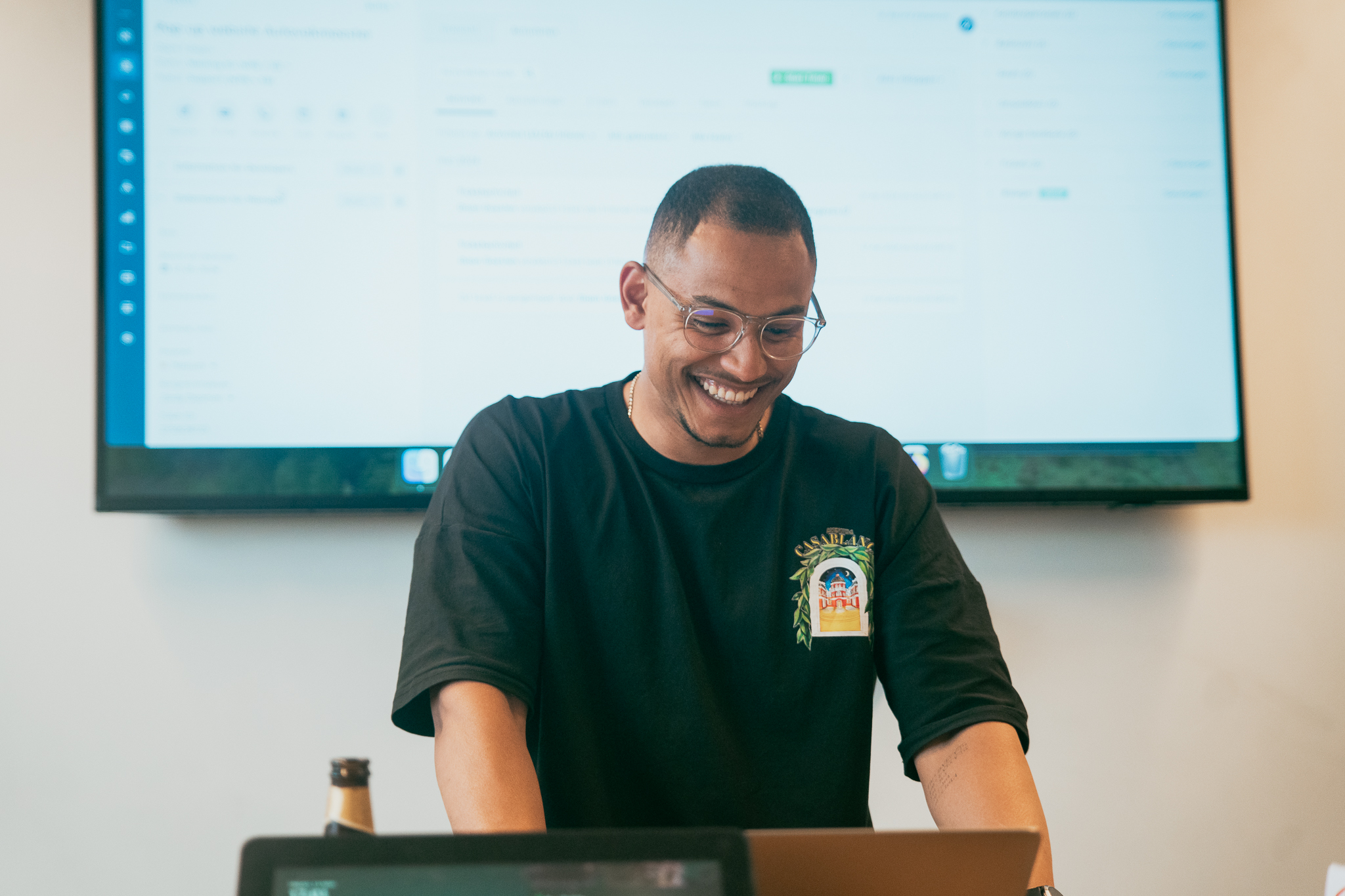 Smiling man in glasses standing at a desk with laptop and large screen behind him.