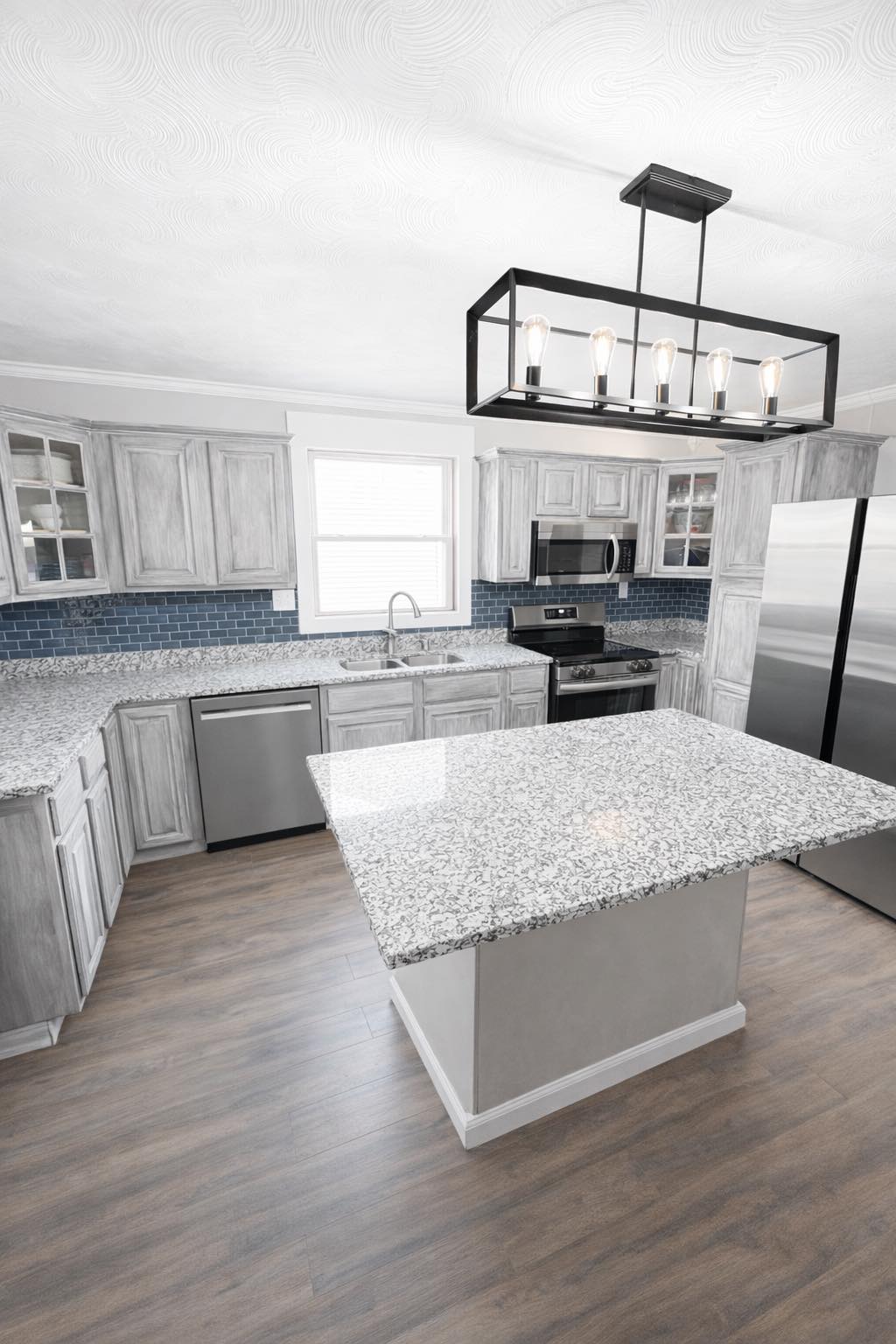 Modern kitchen with a large island, gray cabinets, stainless steel appliances, and dark blue tile backsplash.