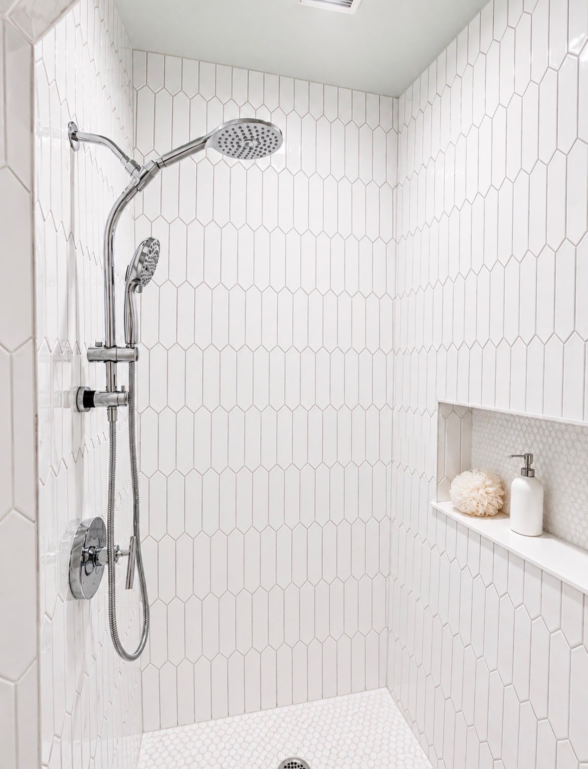 Shower with white geometric tiles, dual showerheads, and a niche holding a loofah and soap dispenser.