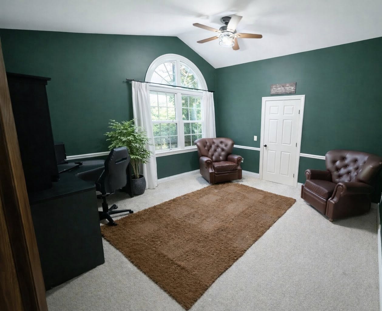 Room with dark green walls, a large window, brown rug, leather chairs, desk, and ceiling fan.
