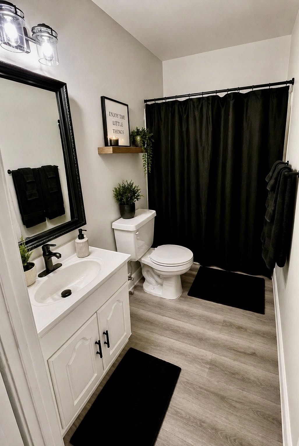 Modern bathroom with white vanity, black fixtures, black shower curtain, rugs, and decorative plants.