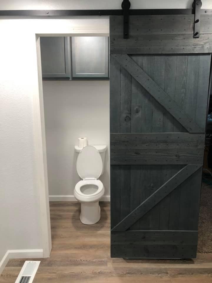 Bathroom with white toilet, two toilet paper rolls, gray cabinets, sliding barn-style door, and wood floor.