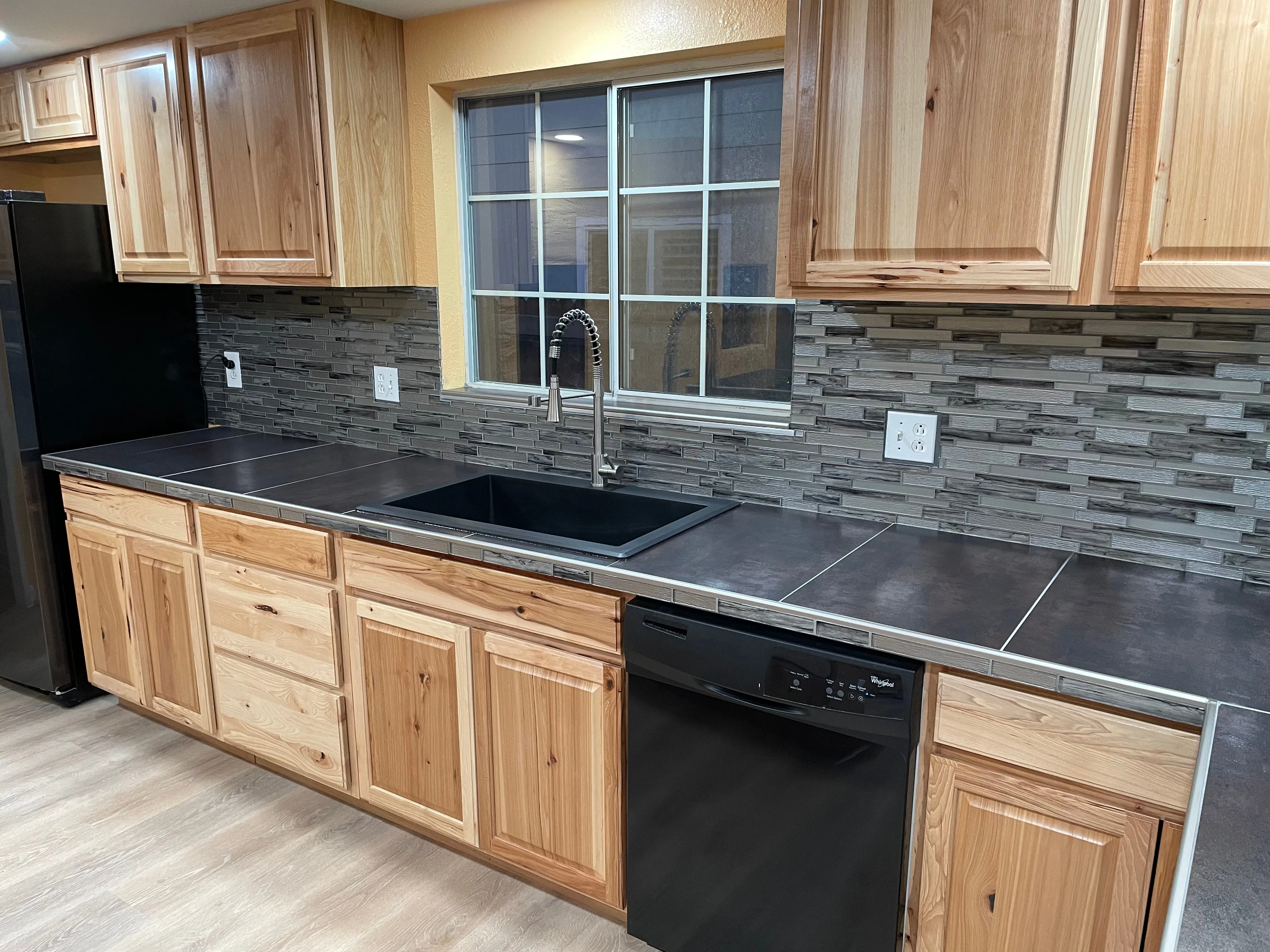 Kitchen with wooden cabinets, black dishwasher, black sink, gray tile countertop, and window above sink.