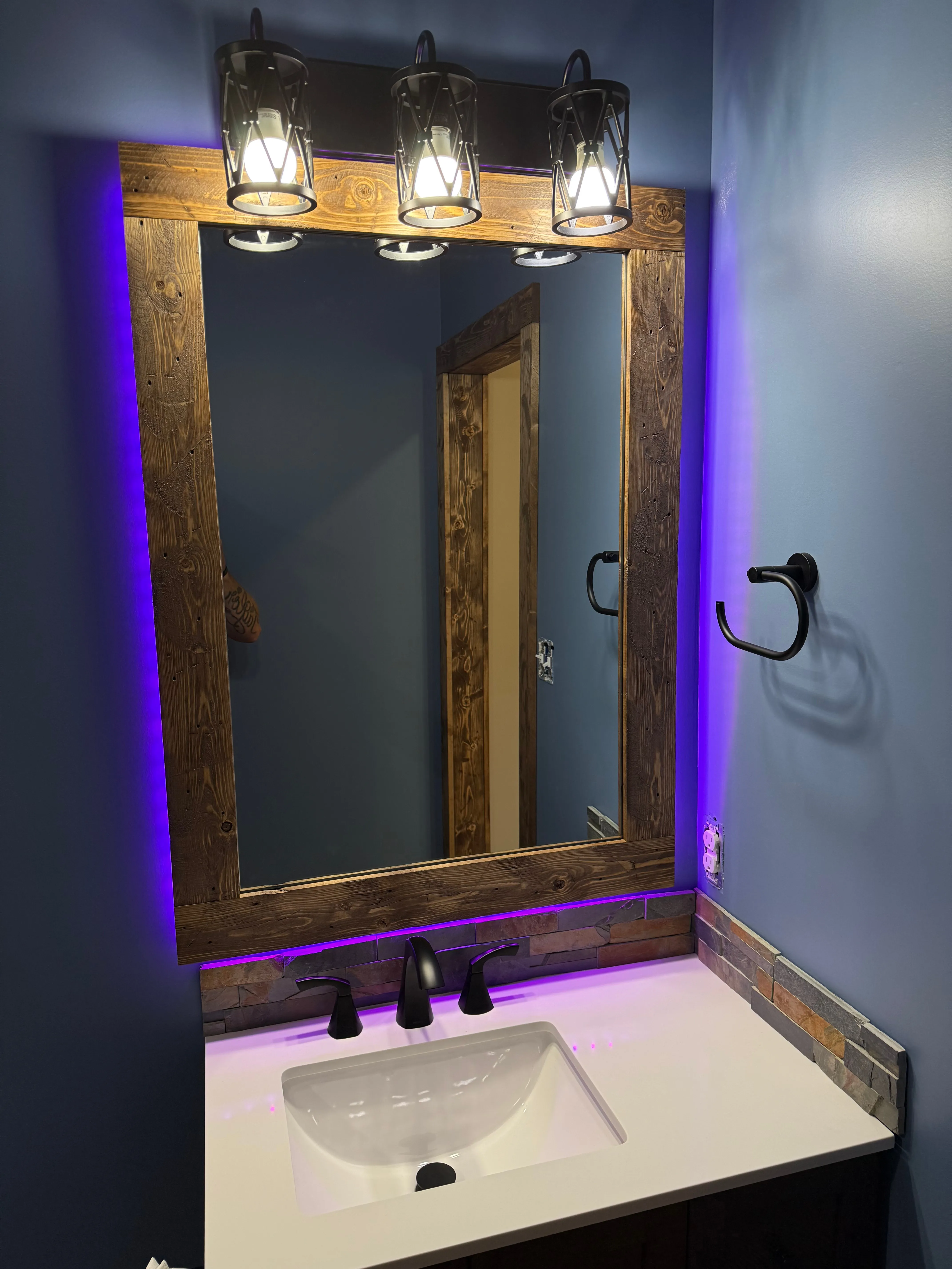 Bathroom vanity with white sink, black faucet, wood-framed mirror, and purple backlighting on blue walls.
