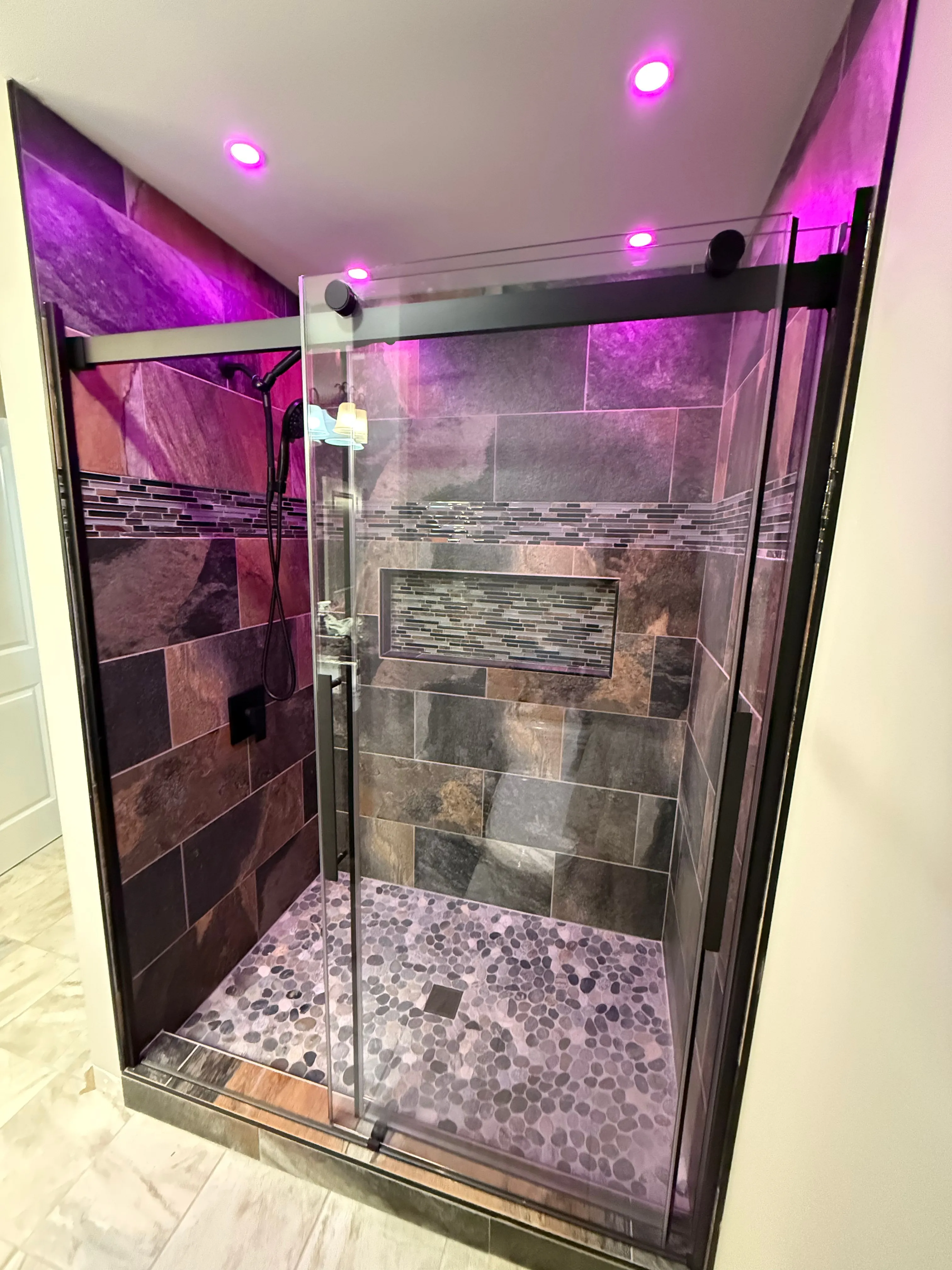 Modern shower with glass door, pebble floor tiles, dark wall tiles, and purple ceiling lights.