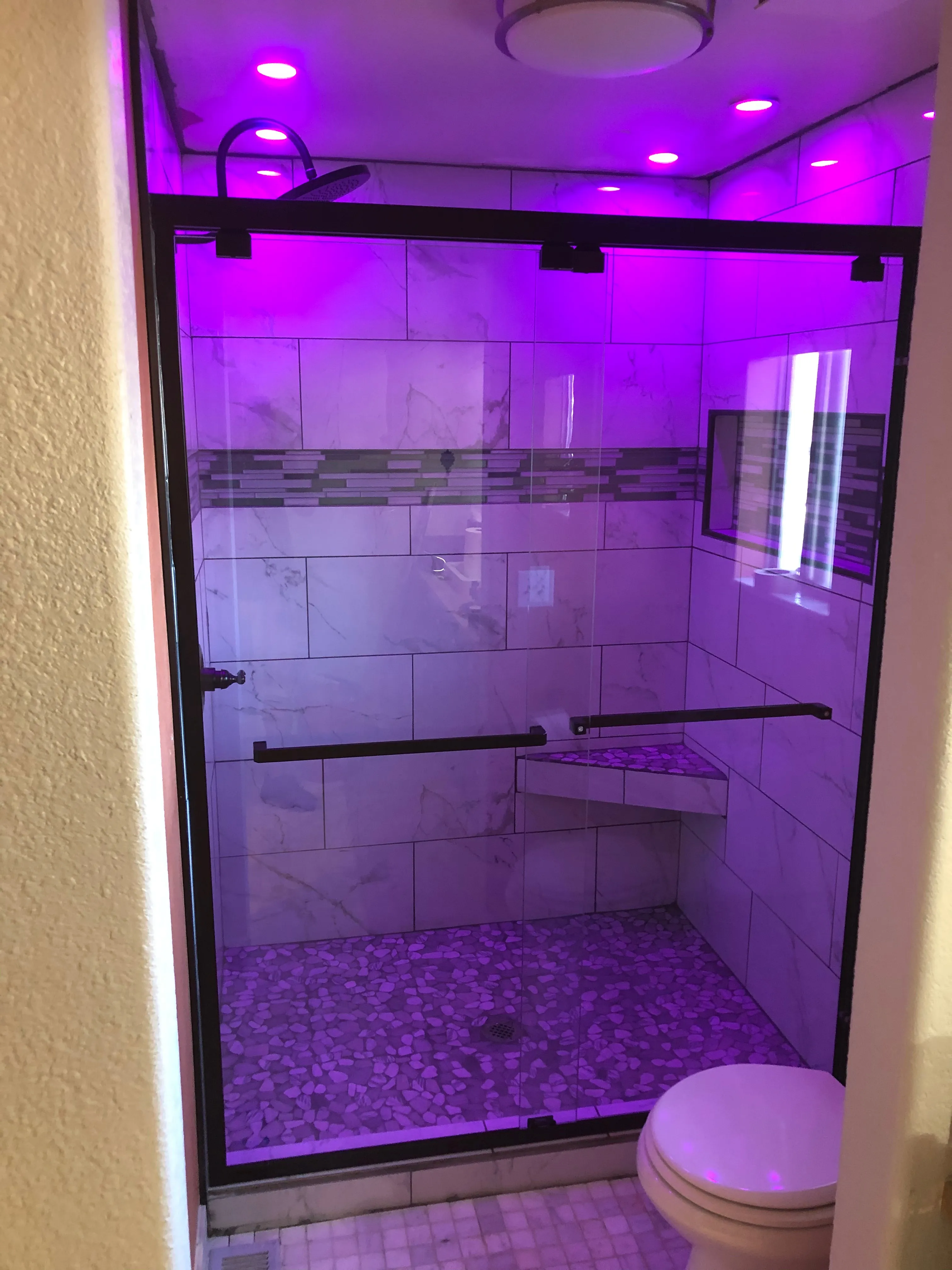 Modern shower with pebble floor, tiled walls, glass door, corner bench, and purple ceiling lights.