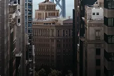 Tall buildings closely packed in a city with a bridge visible in the background under a cloudy sky.
