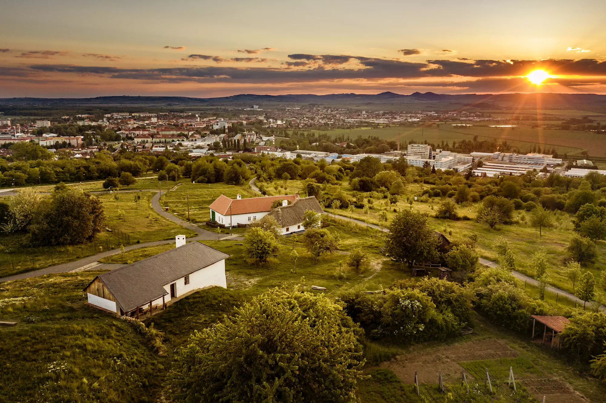 Západ slunce nad zelenými poli s menšími bílými domy a vzdáleným Uherským Hradištěm na horizontu kopcovité krajiny.