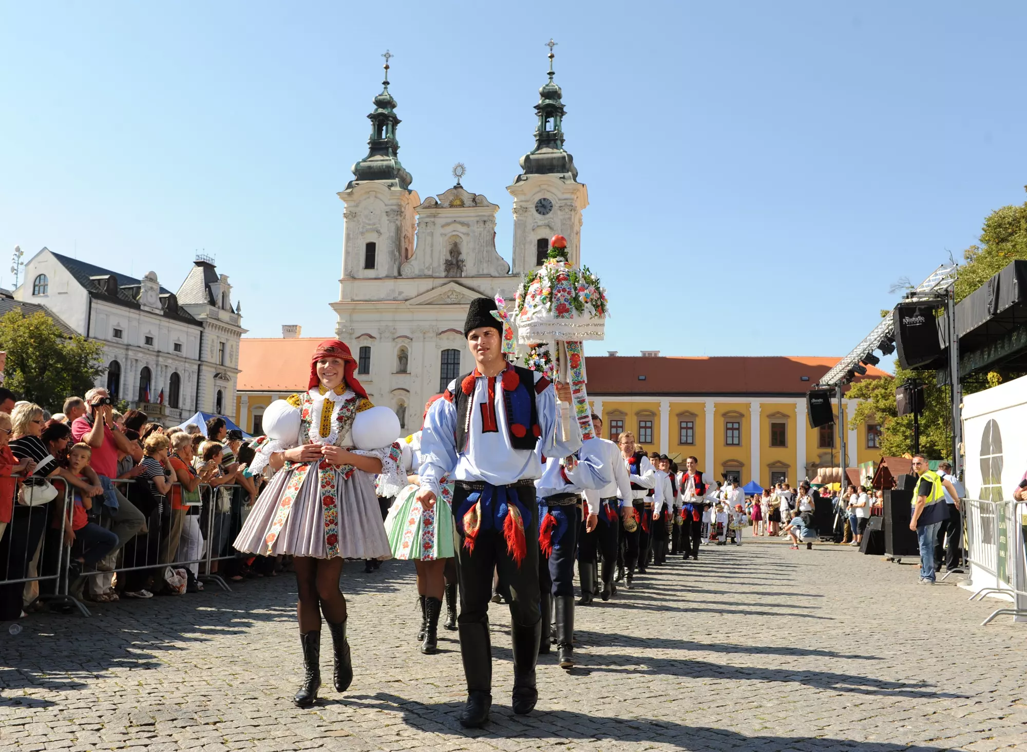Průvod lidí v tradičních lidových krojích prochází dlážděnou ulicí kolem historického kostela v Uherském Hradišti.