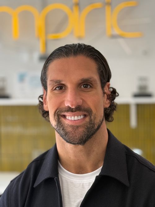Smiling man with dark hair and beard wearing black jacket over white shirt in indoor setting.