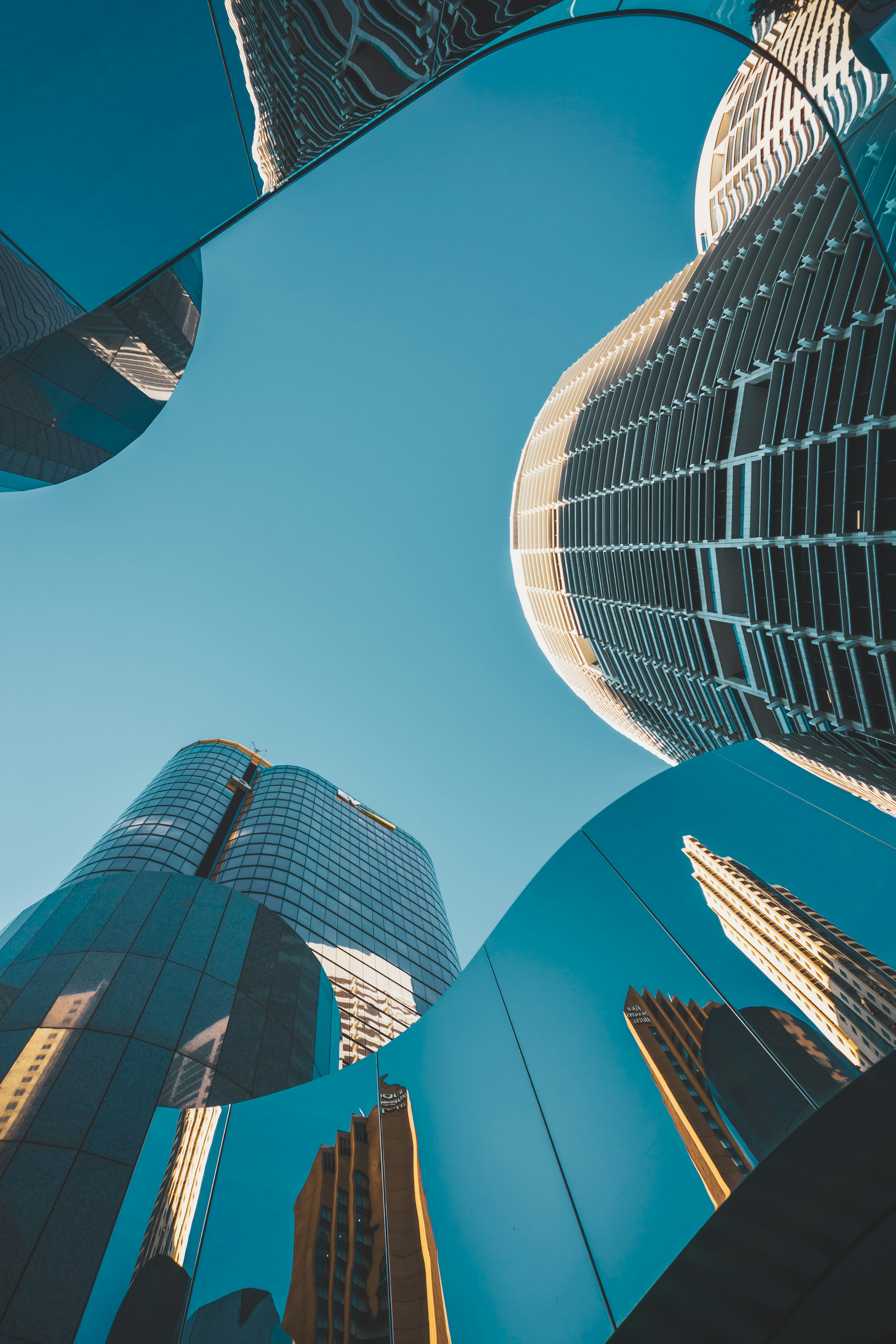 Upward view of modern skyscrapers with reflections in curved glass panels against blue sky.