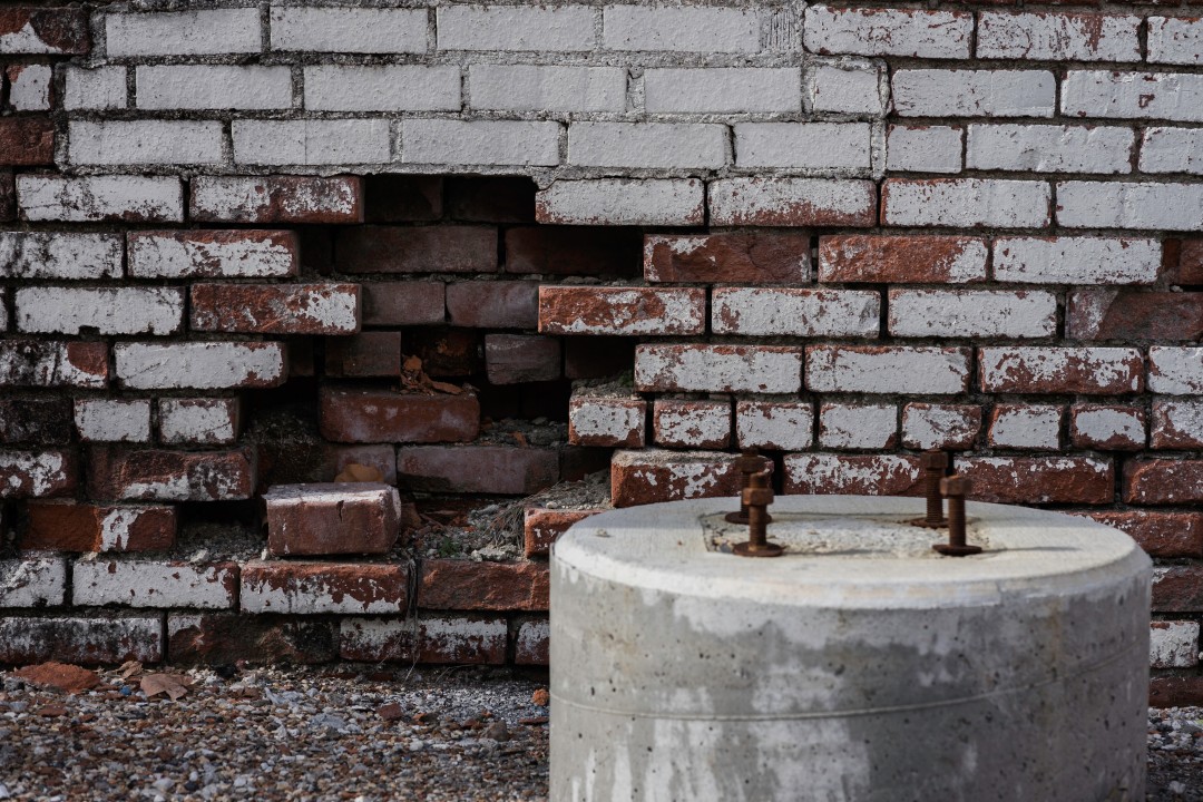 Old brick wall with a large hole and a concrete cylinder with rusted bolts in front.