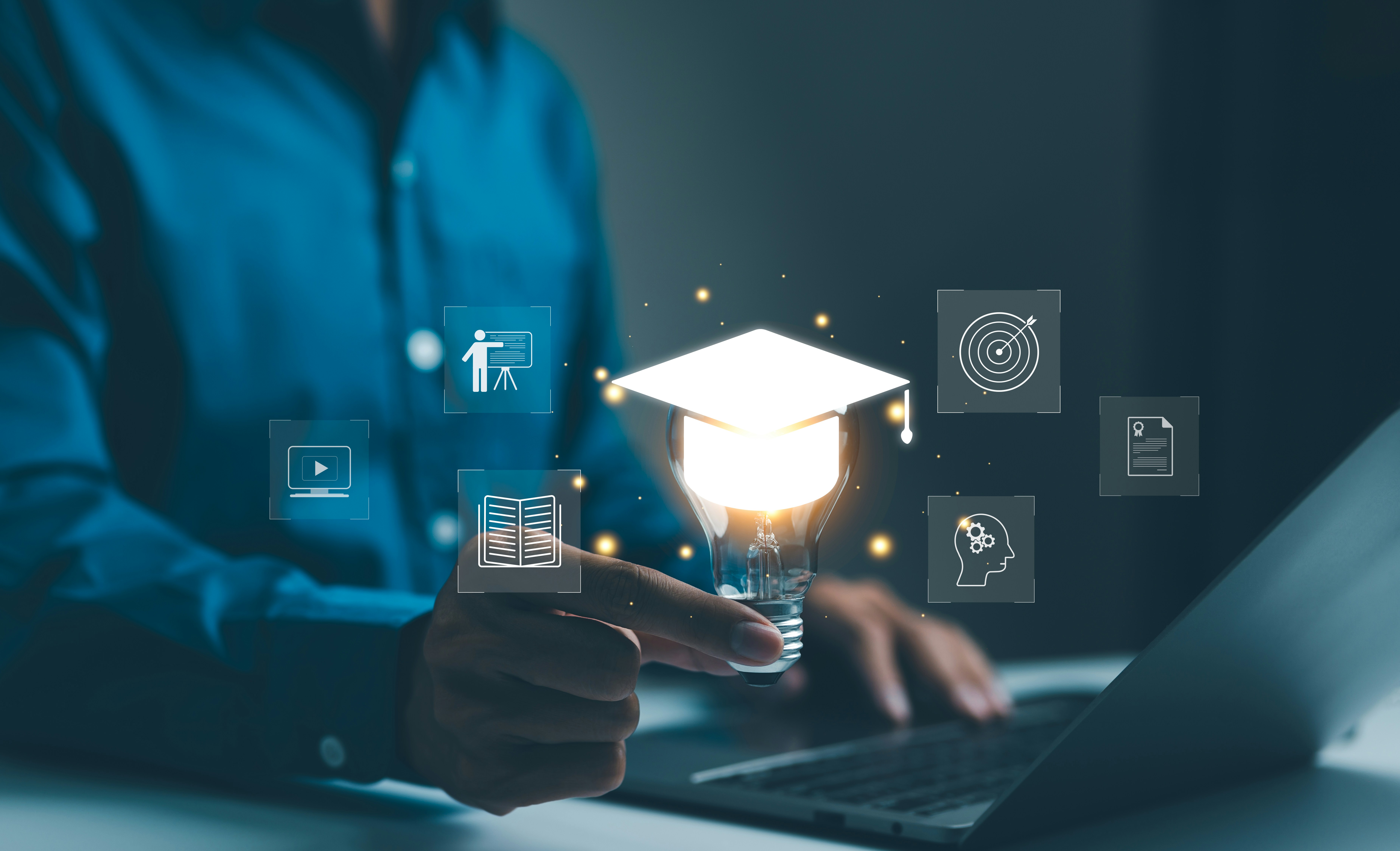 Hand holding lightbulb with glowing graduation cap icon surrounded by education-related symbols by laptop.