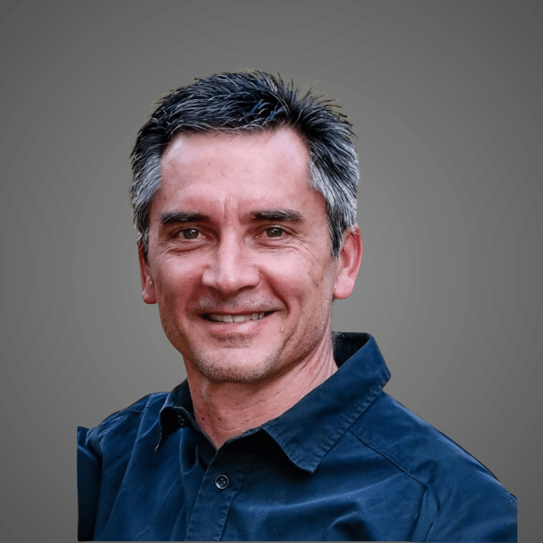 Smiling middle-aged man with salt-and-pepper hair wearing a dark blue collared shirt on a gray background.