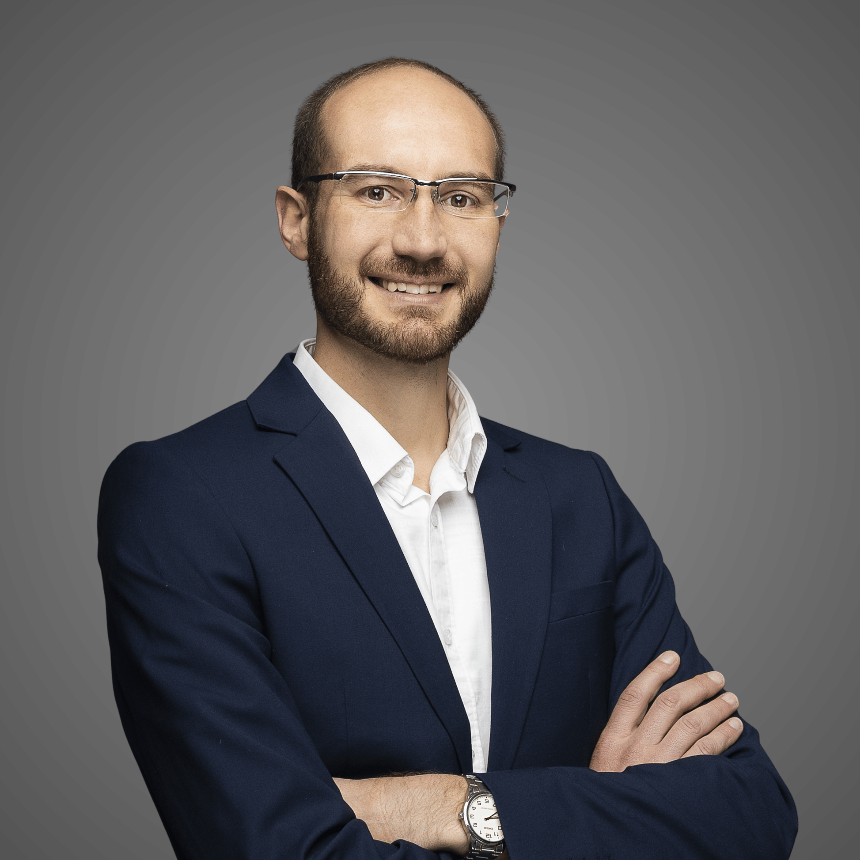 Smiling man with glasses in a navy blazer and white shirt with arms crossed against a gray background.