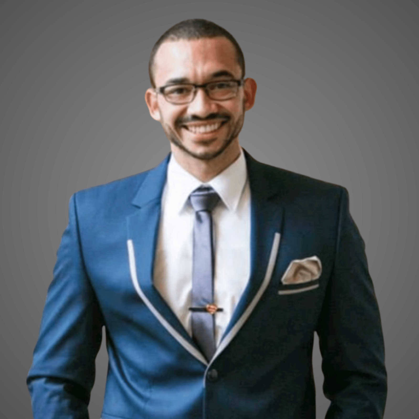 Smiling man wearing glasses and a blue suit with a white shirt and tie on a gray background.