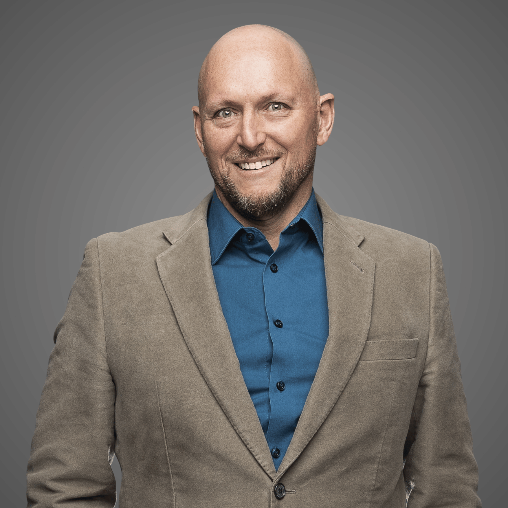 Smiling bald man with beard wearing blue shirt and tan blazer against gray background.