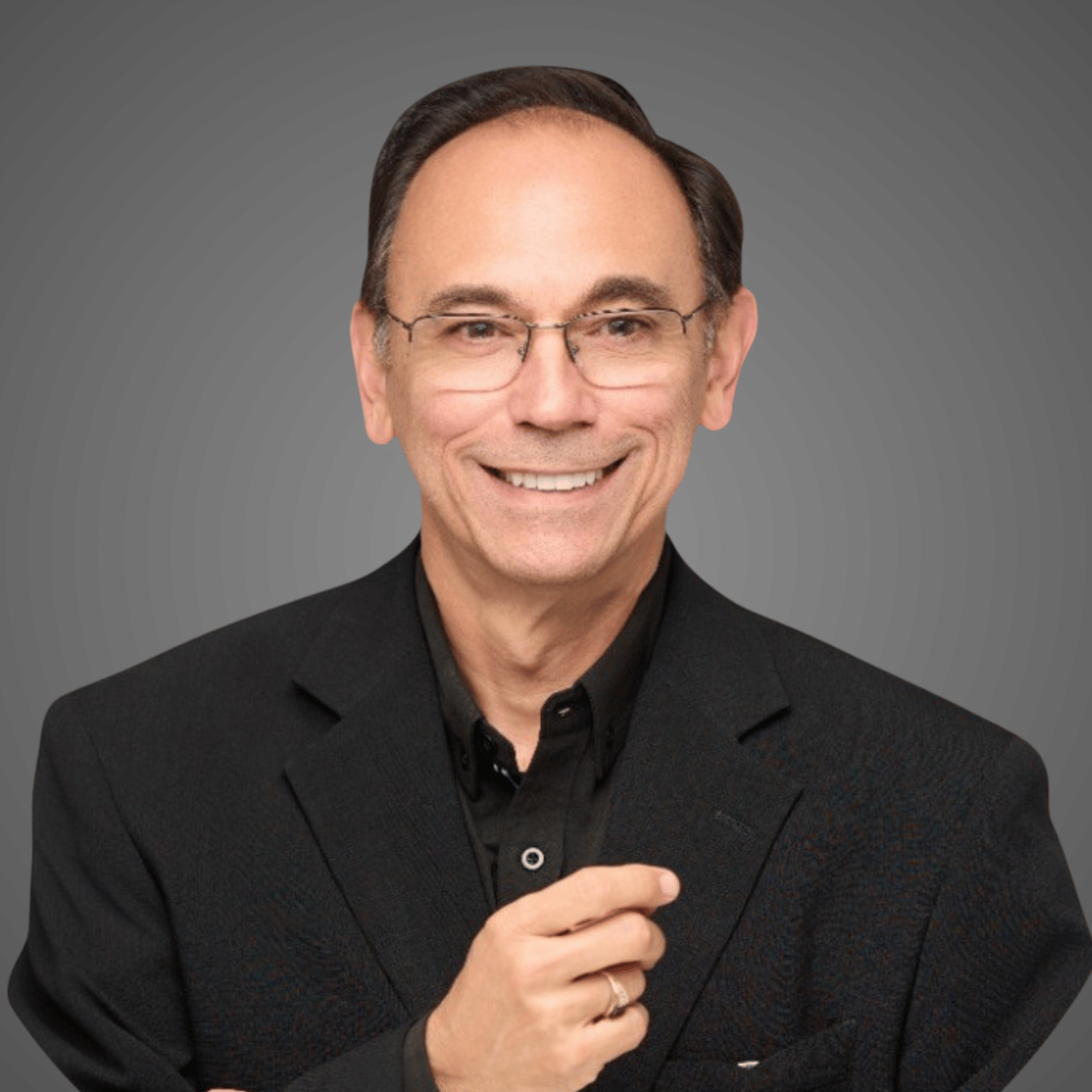 Smiling man with glasses wearing a black suit and shirt, posing against a gray background.