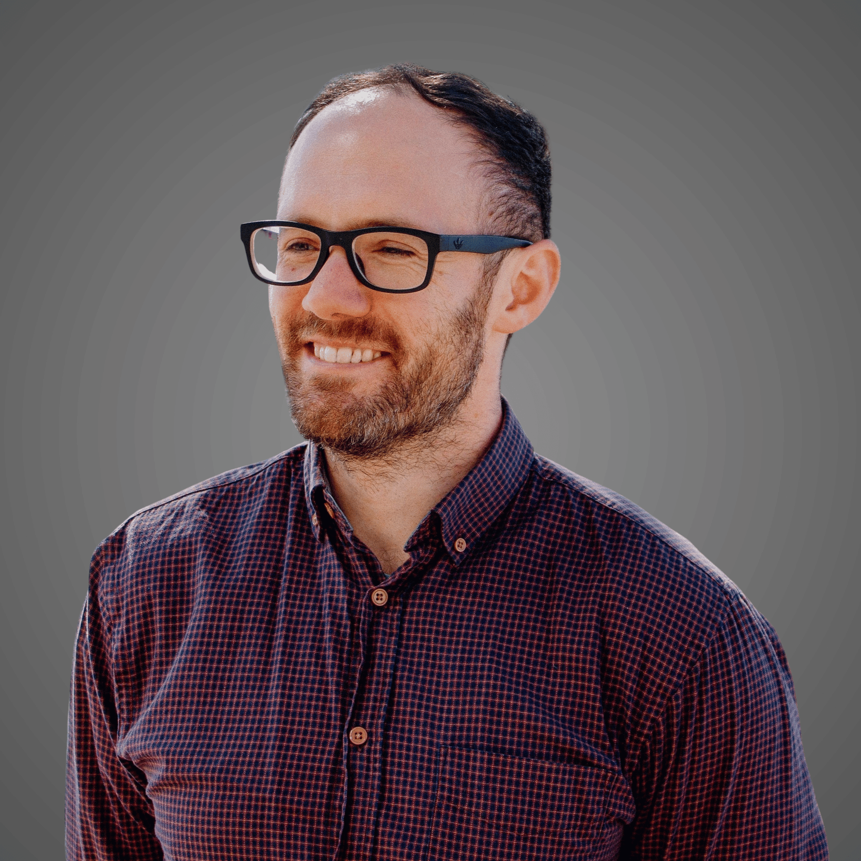 Smiling man with glasses and beard wearing a checkered button-up shirt on gray background.