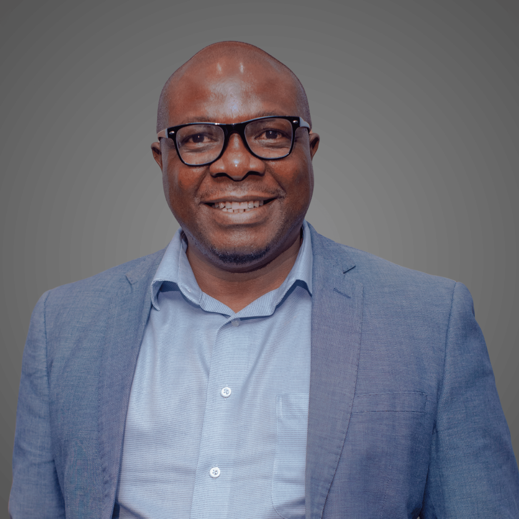 Smiling man wearing glasses, light blue shirt, and blue blazer in front of a gray background.