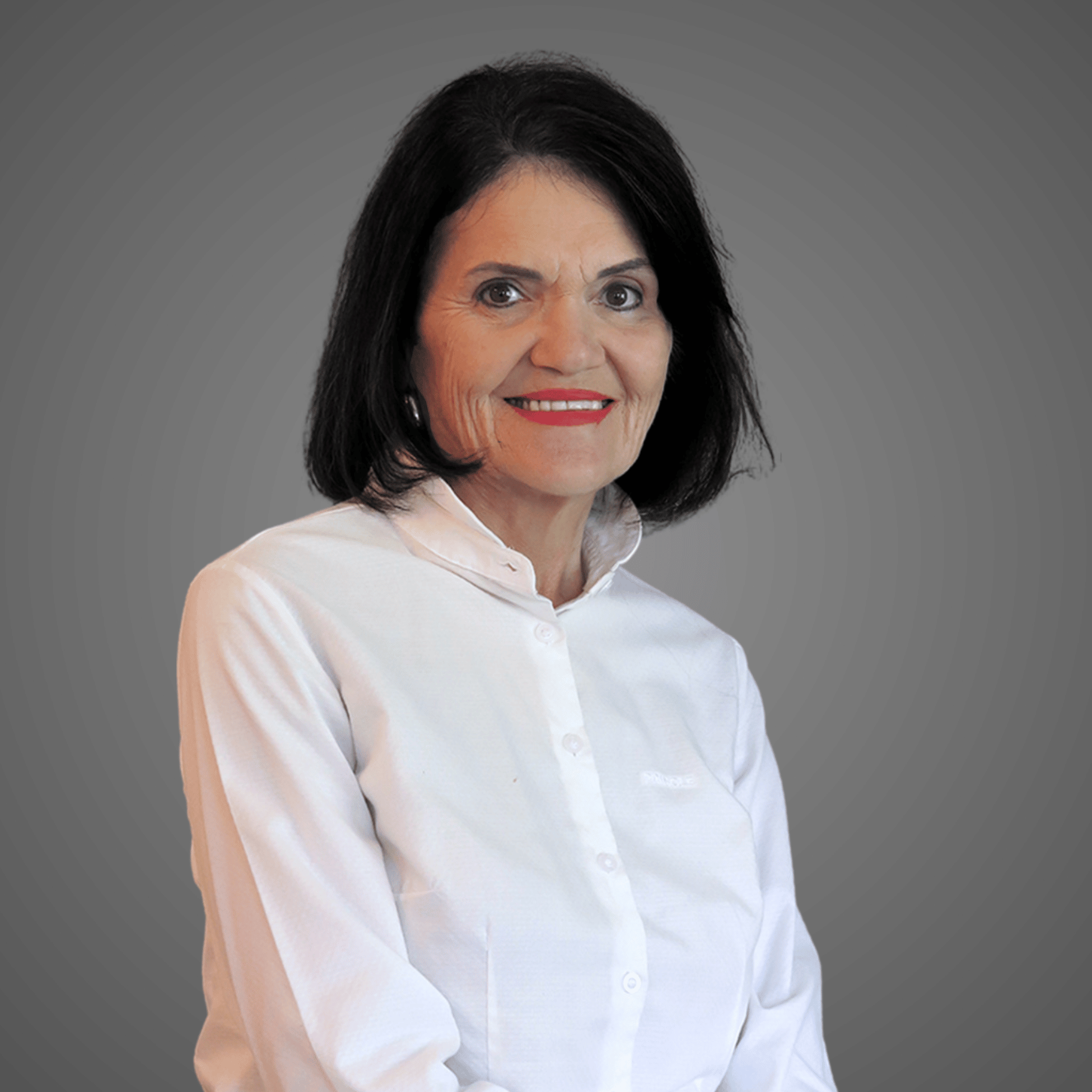 Smiling older woman with dark hair wearing a white buttoned shirt against a gray background.