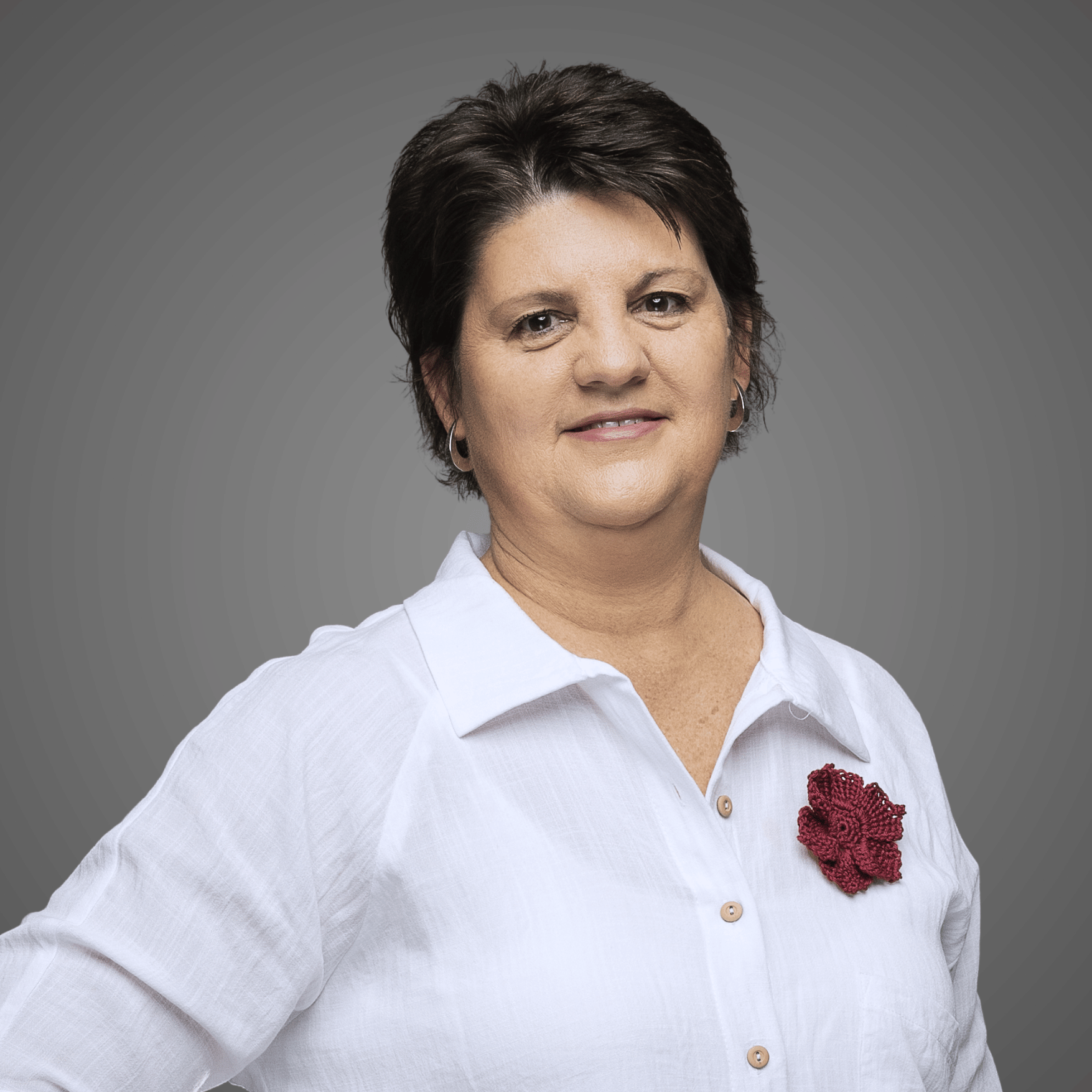 Woman with short dark hair wearing white shirt with a red crocheted flower pin on a gray background.