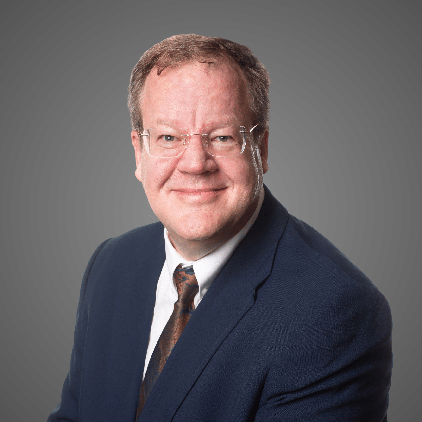 Smiling man in glasses wearing a navy suit jacket, white shirt, and dark patterned tie on gray background.