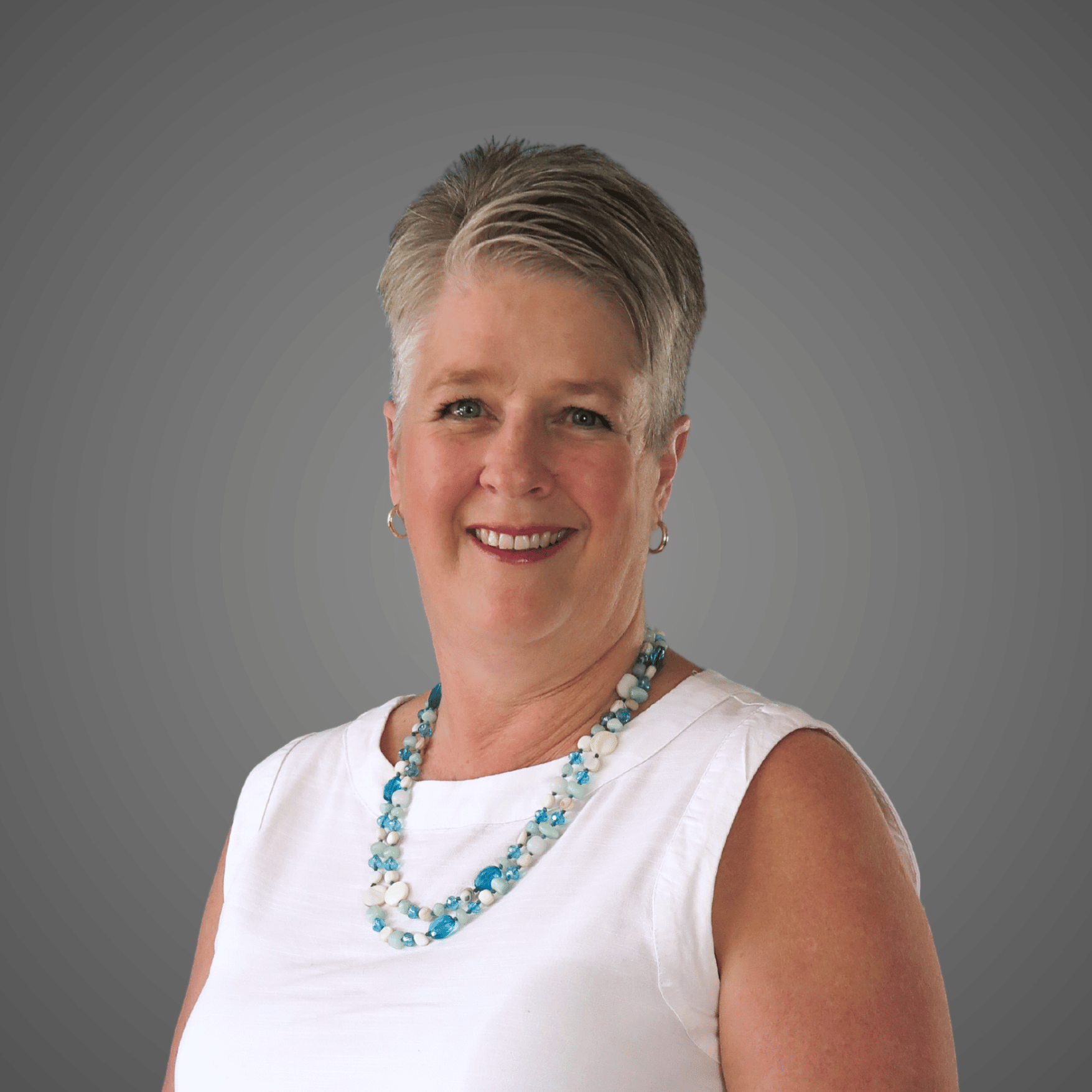 Smiling woman with short gray hair wearing white sleeveless top and blue-white beaded necklace.