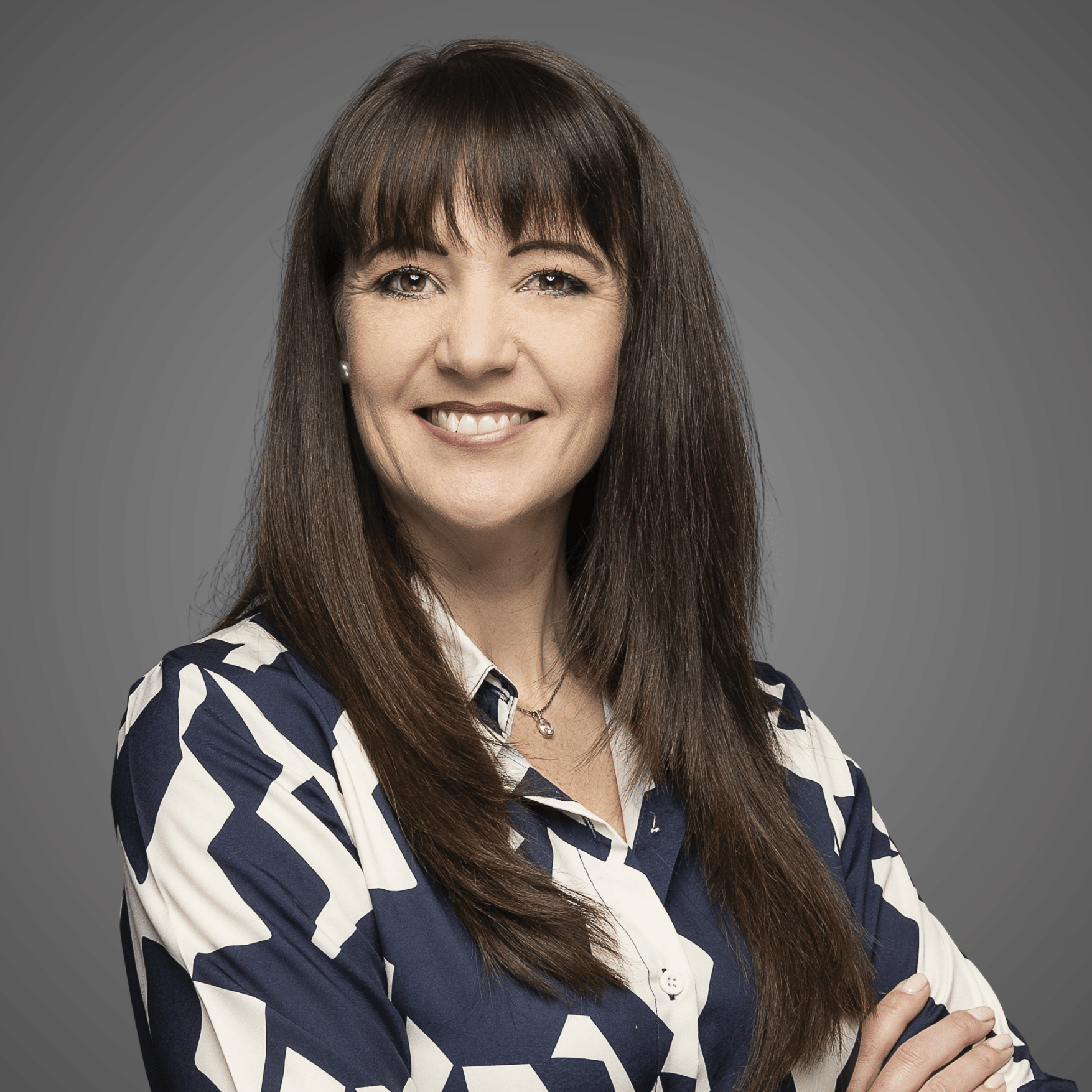 Smiling woman with long brown hair wearing a patterned blue and white shirt against a gray background.