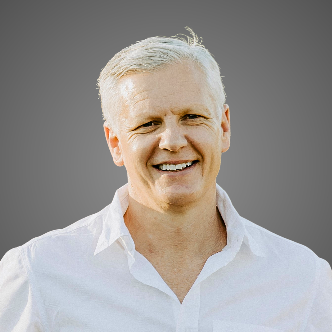 Smiling middle-aged man with short white hair wearing a white button-up shirt on gray background.