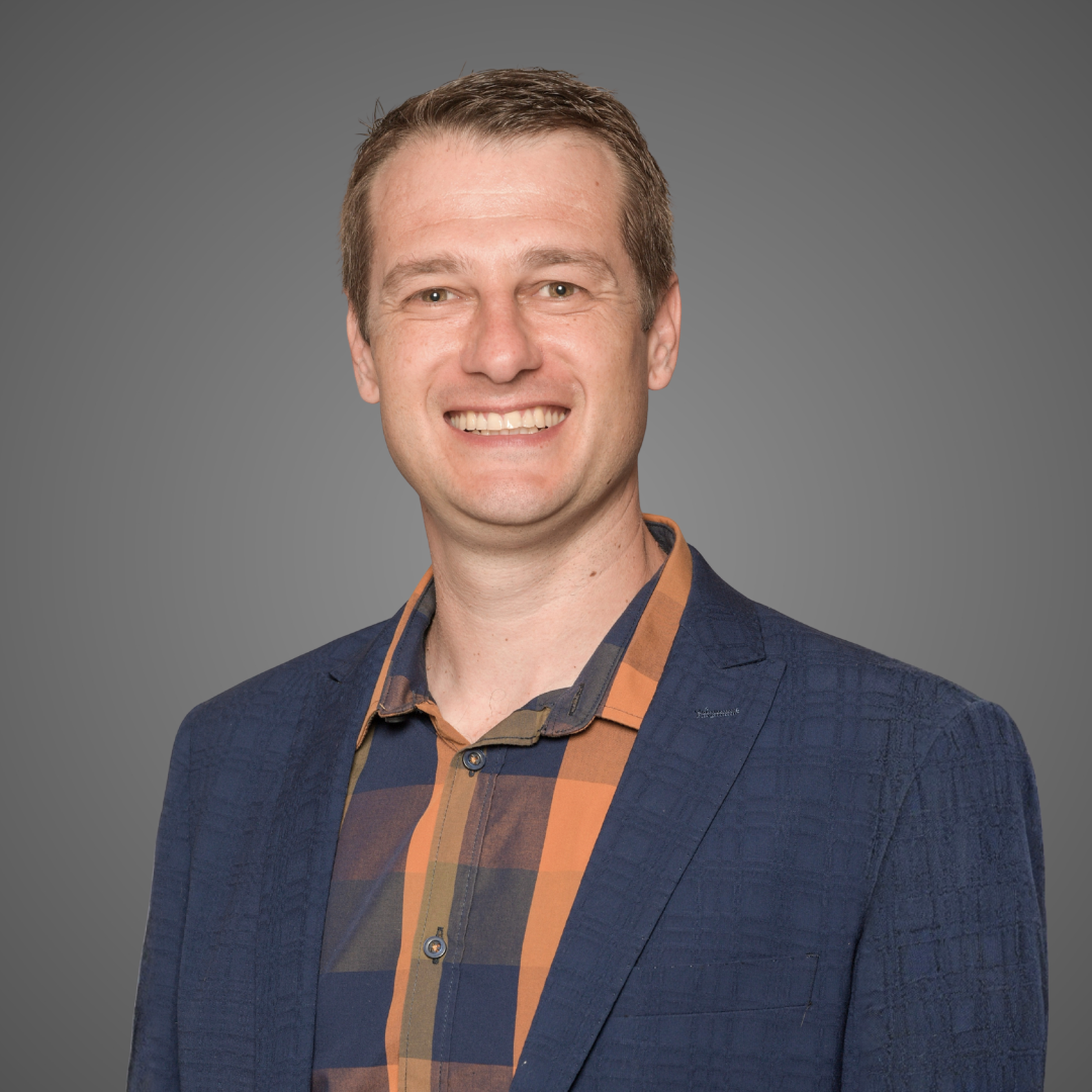 Smiling man in a blue blazer and orange plaid shirt against a gray gradient background.