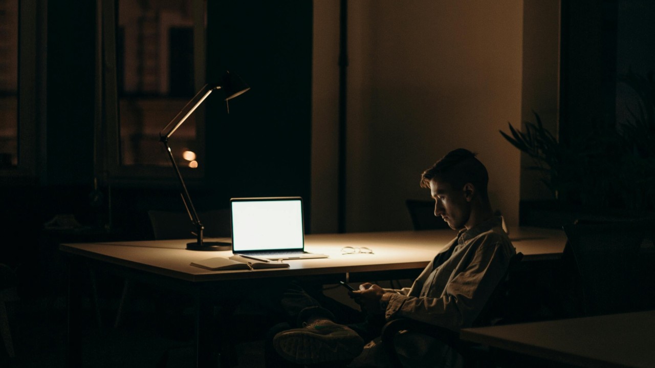 Man sitting alone in dimly lit office at night, looking at his phone beside a laptop on the desk.