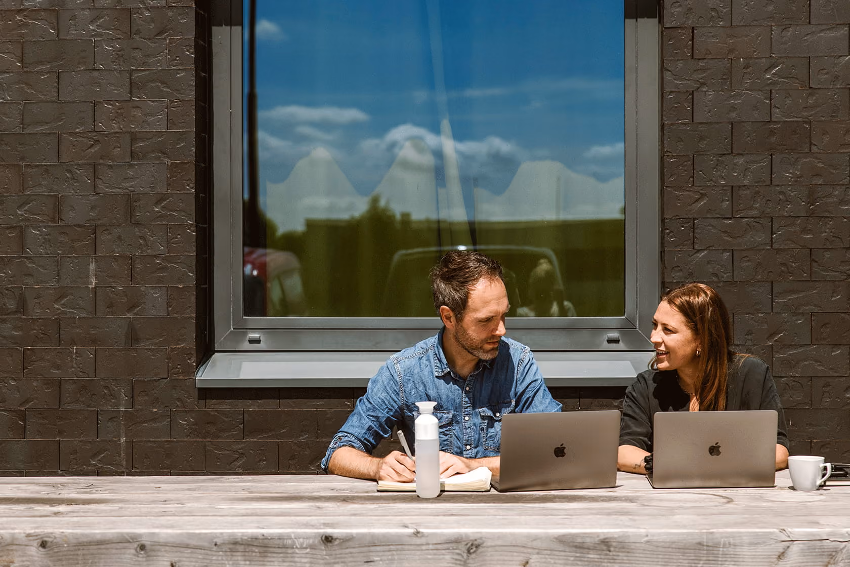 Man en vrouw werken buiten aan laptops bij een houten tafel onder een raam op een zonnige dag.