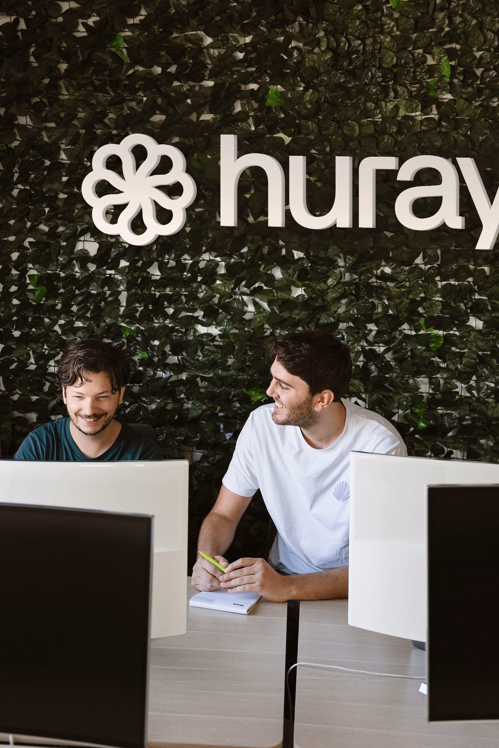 Twee mannen praten en lachen bij een bureau met computers voor een groene muur met het woord huray.