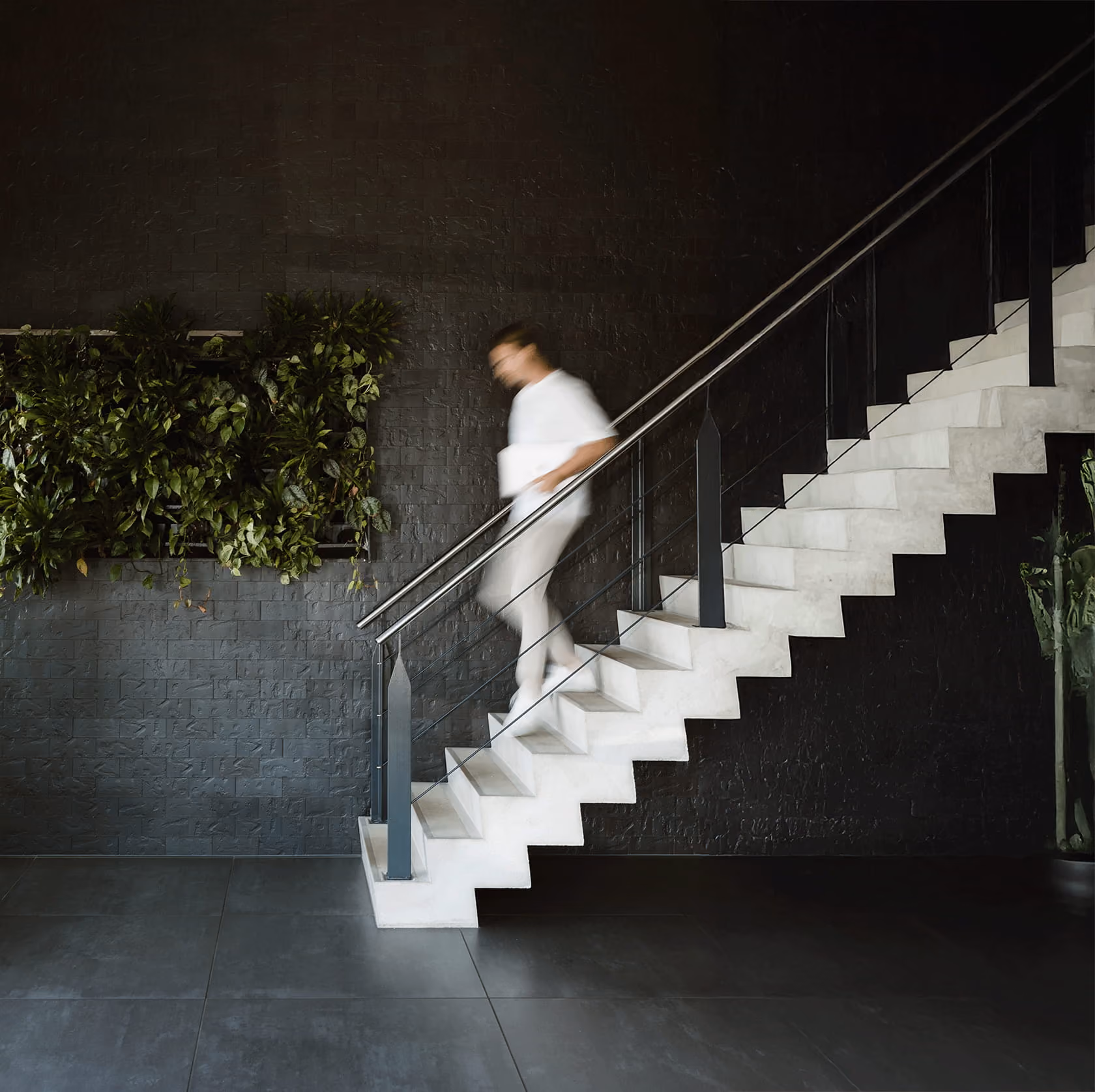 Bewegende persoon in witte kleding daalt een trap af in een moderne ruimte met groene planten aan de muur.