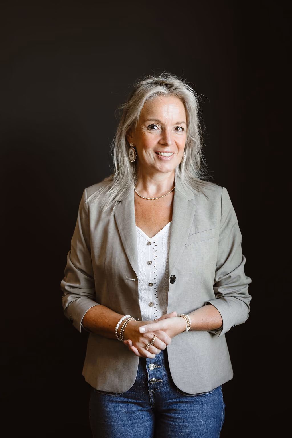 Blonde vrouw in beige blazer en jeans lacht vriendelijk tegen donkere achtergrond.