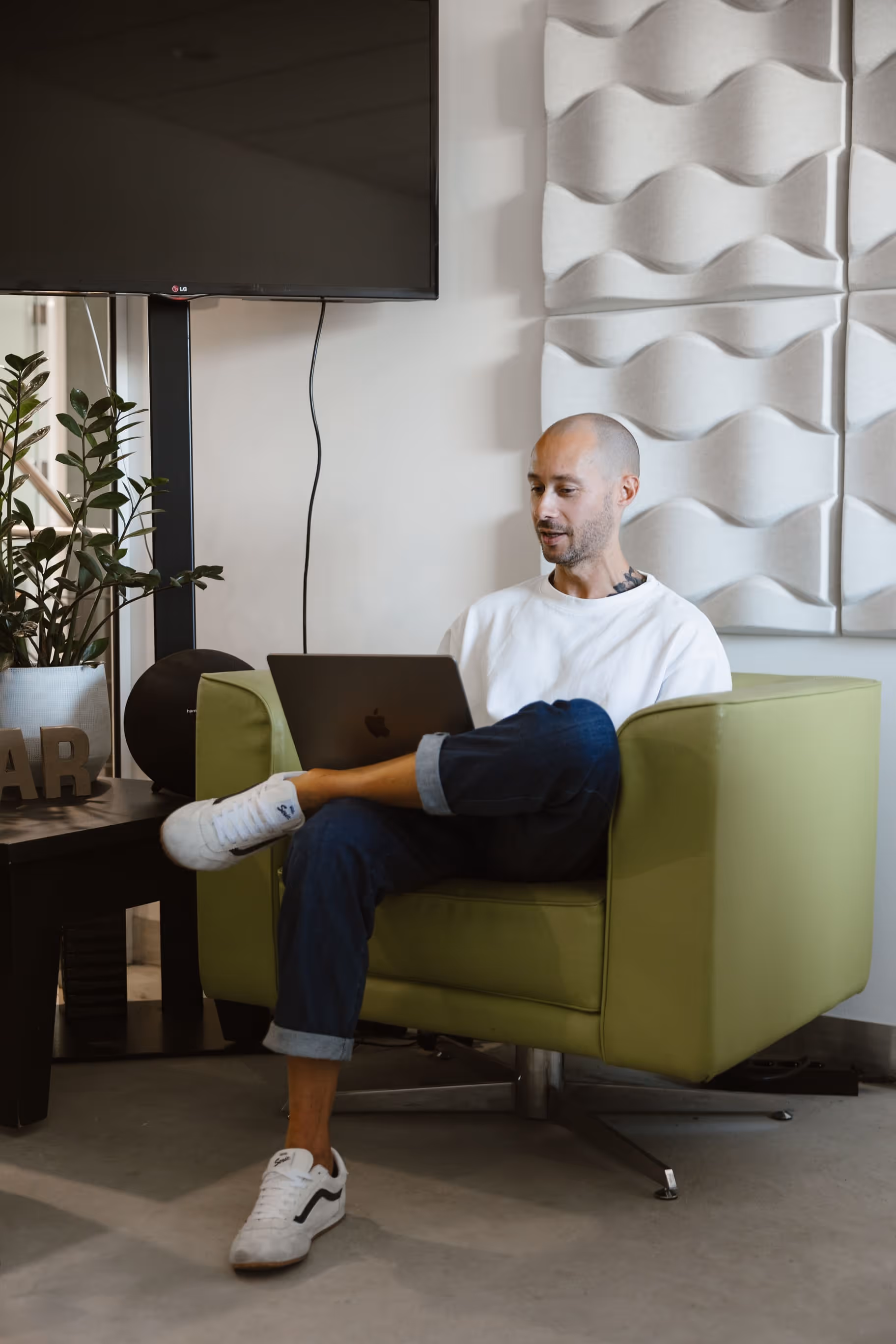 Man met witte trui en sneakers werkt op laptop in een groene stoel in een moderne kamer.
