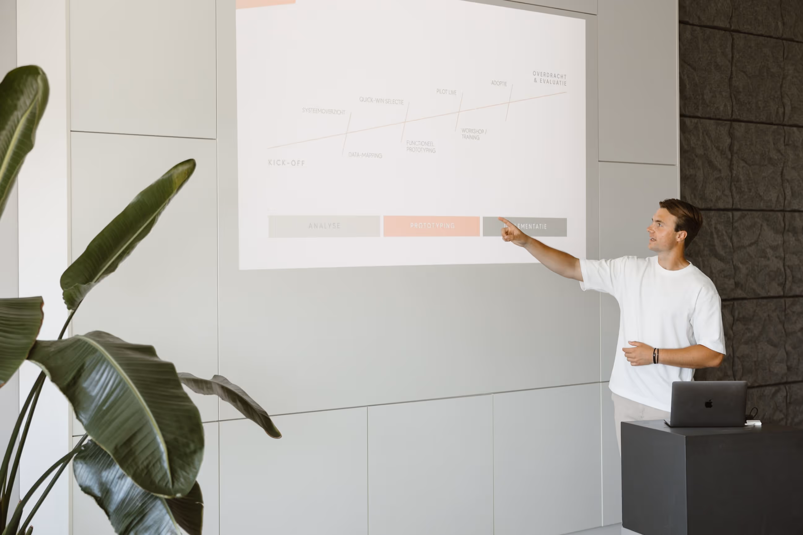 Man wijst naar een projectstroomdiagram op een muur terwijl hij voor een laptop staat in een vergaderruimte.